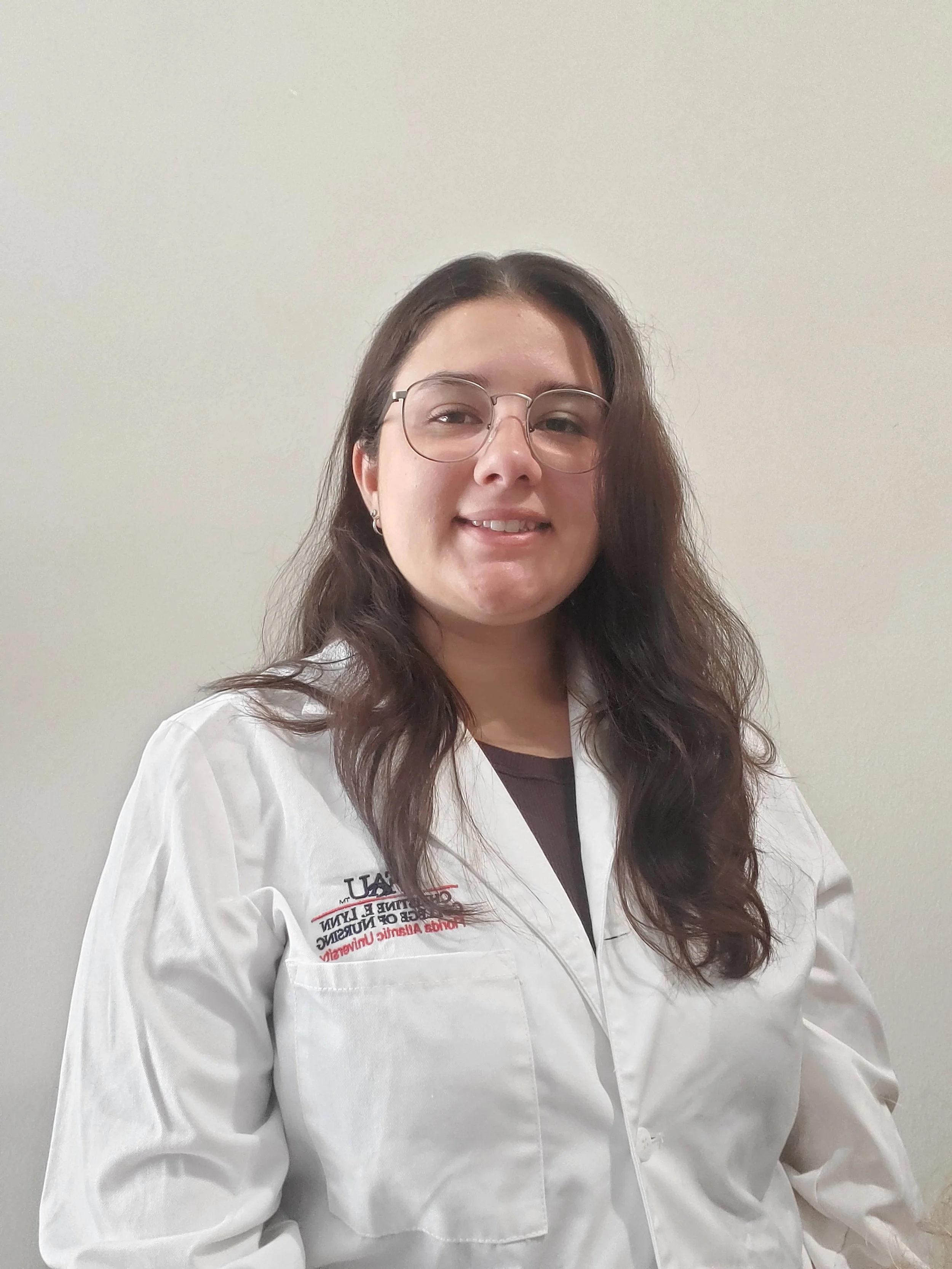 A young woman with long wavy brown hair, glasses, and earrings wearing a white medical coat standing against a plain light-colored wall.