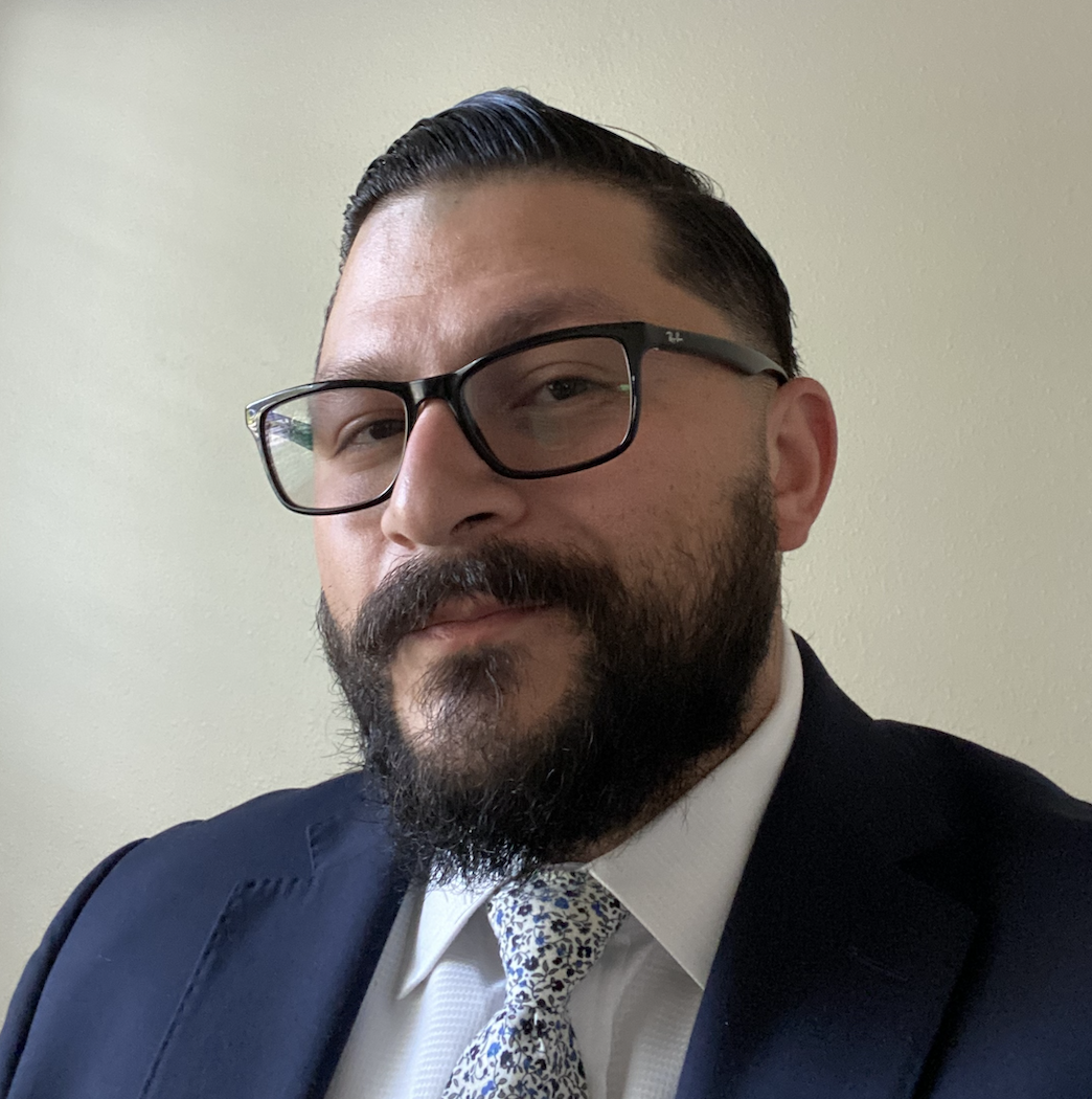 A man with glasses, a beard, and neatly combed hair, dressed in a suit and tie, posing against a plain wall.
