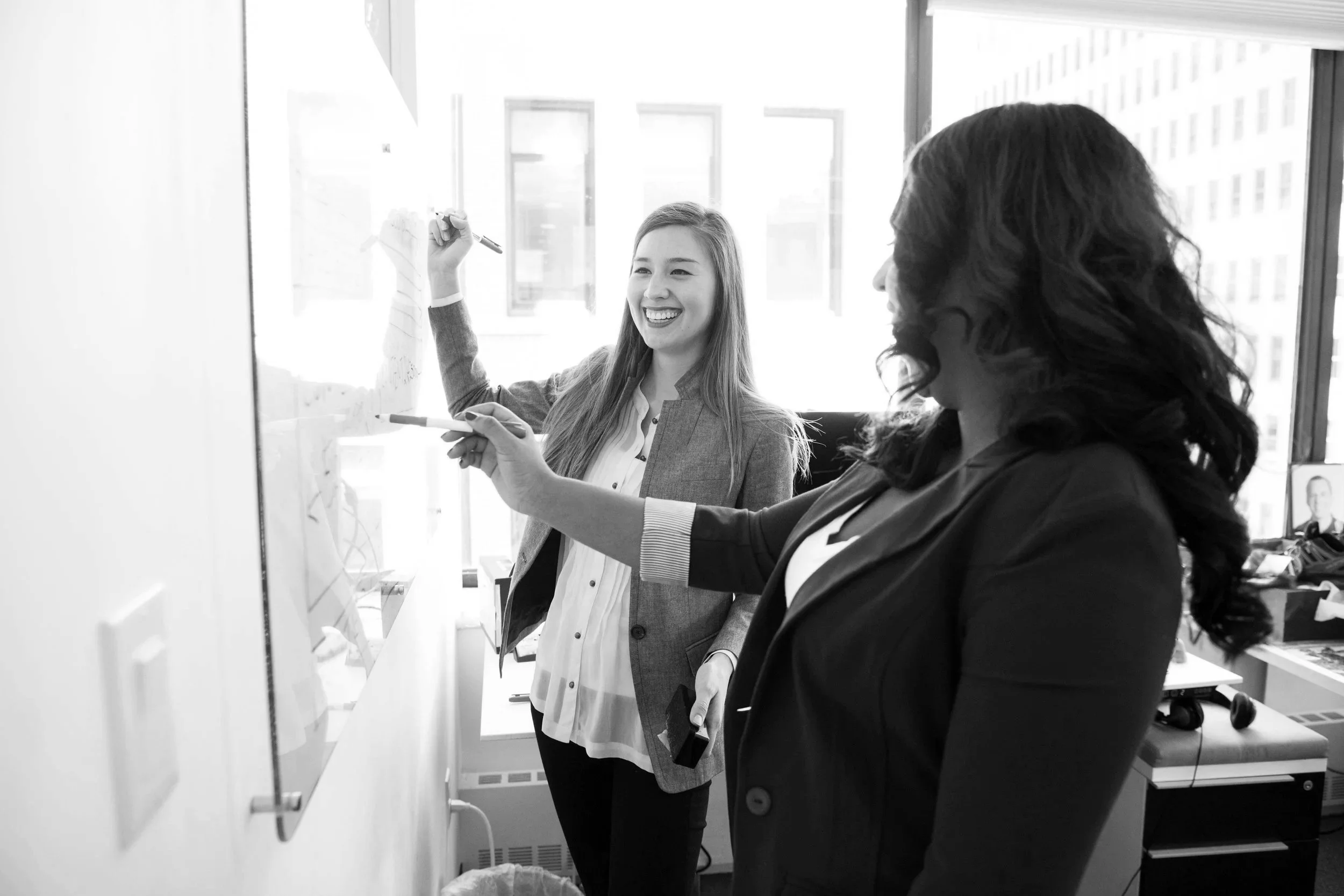 Two women at Wednesday Ventures discussing a project in the office, one writing on a whiteboard and the other observing, with large windows in the background.