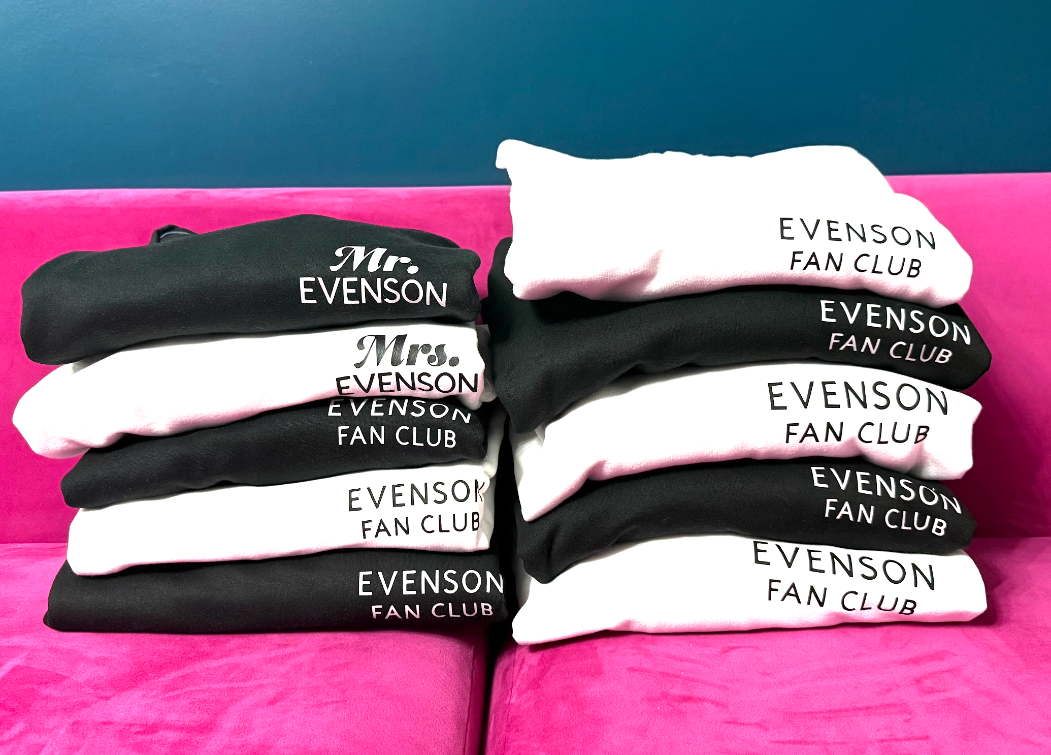 Stacks of black and white sweatshirts with printed text, arranged on a pink surface with a blue wall in the background.