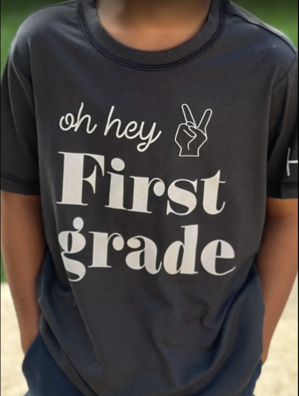 A person wearing a black T-shirt with white text that says 'oh hey First grade' and a hand making a peace sign.