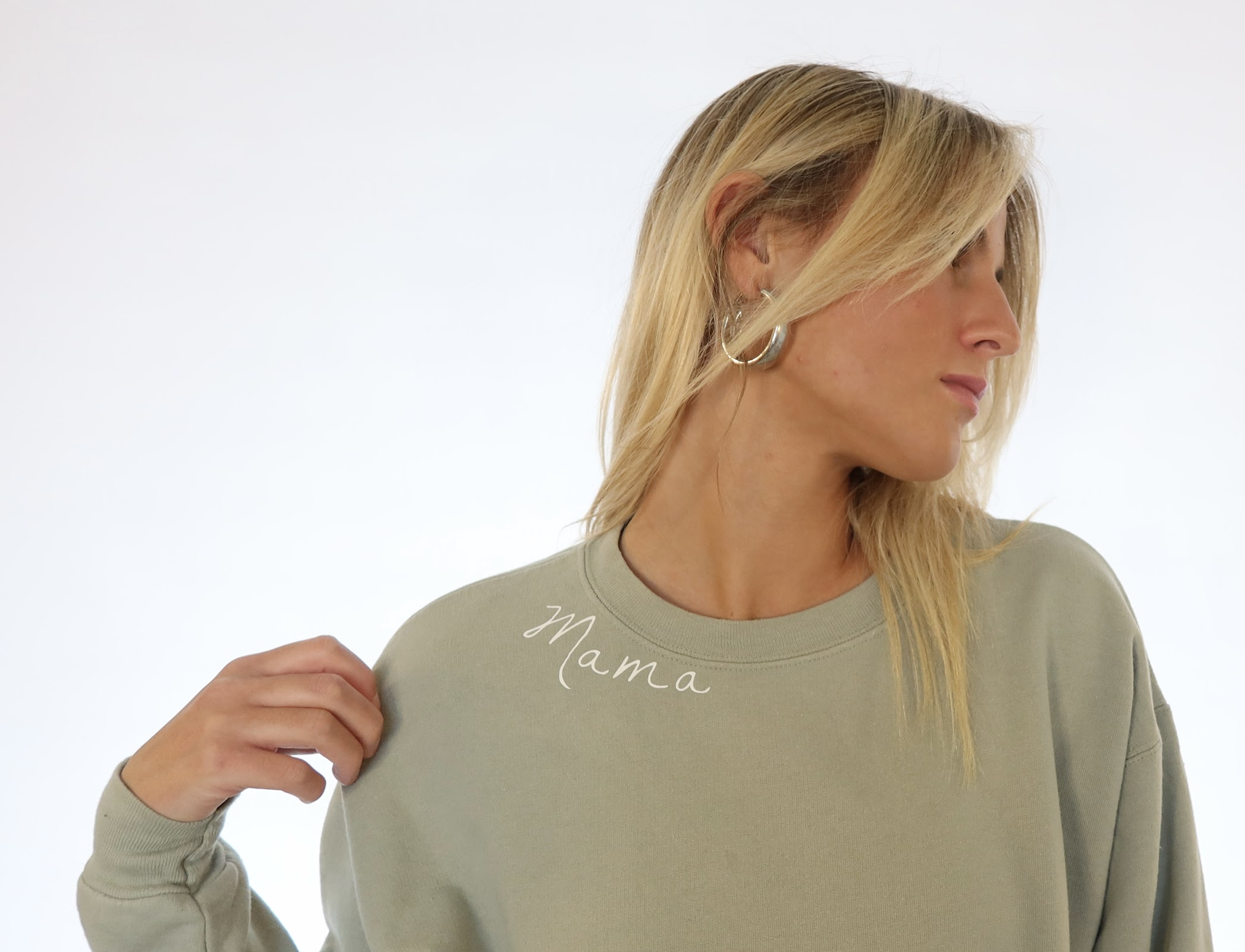 Woman with blonde hair wearing a light gray sweatshirt with 'Mama' embroidered on the collar, holding the shoulder of her sweatshirt, facing sideways with a neutral expression.