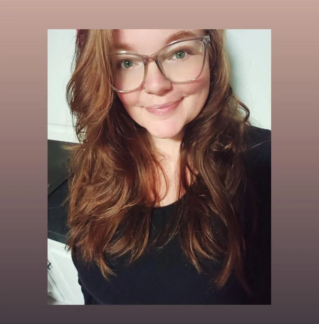 A woman with long, wavy brown hair, glasses, light skin, and a nose piercing, smiling at the camera, wearing a black top.