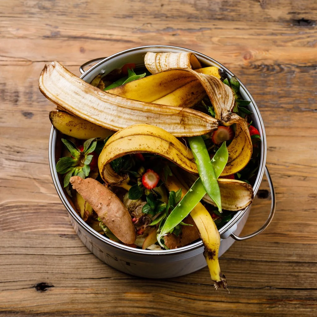 A metal bucket filled with overripe banana peels, strawberry greens, ginger root, and other vegetable scraps, sitting on a wooden surface.