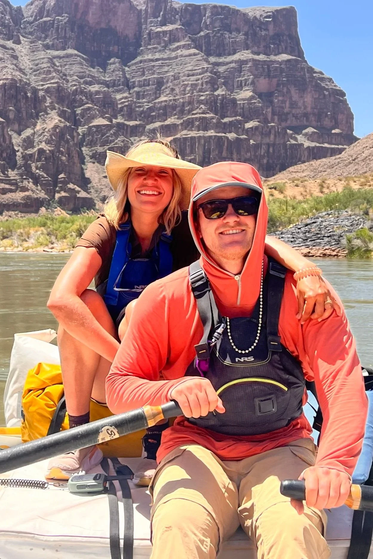 A smiling woman wearing a wide-brimmed hat and a man wearing sunglasses and a hooded jacket paddle boating on a river with Grand Canyon in the background.