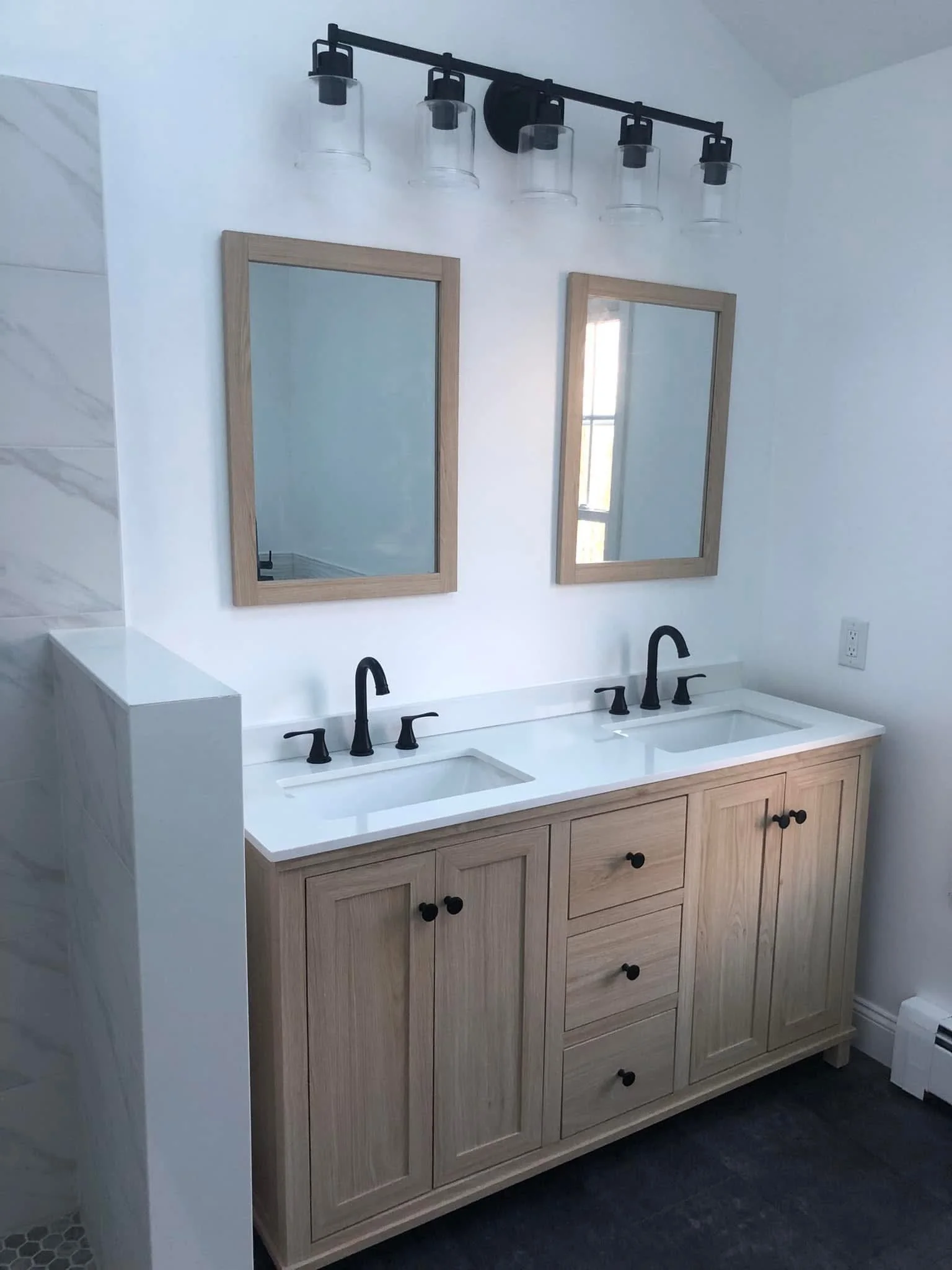 Double vanity bathroom with light wood cabinets, two mirrors with wooden frames, black faucets, and a three-light fixture above mirrors.