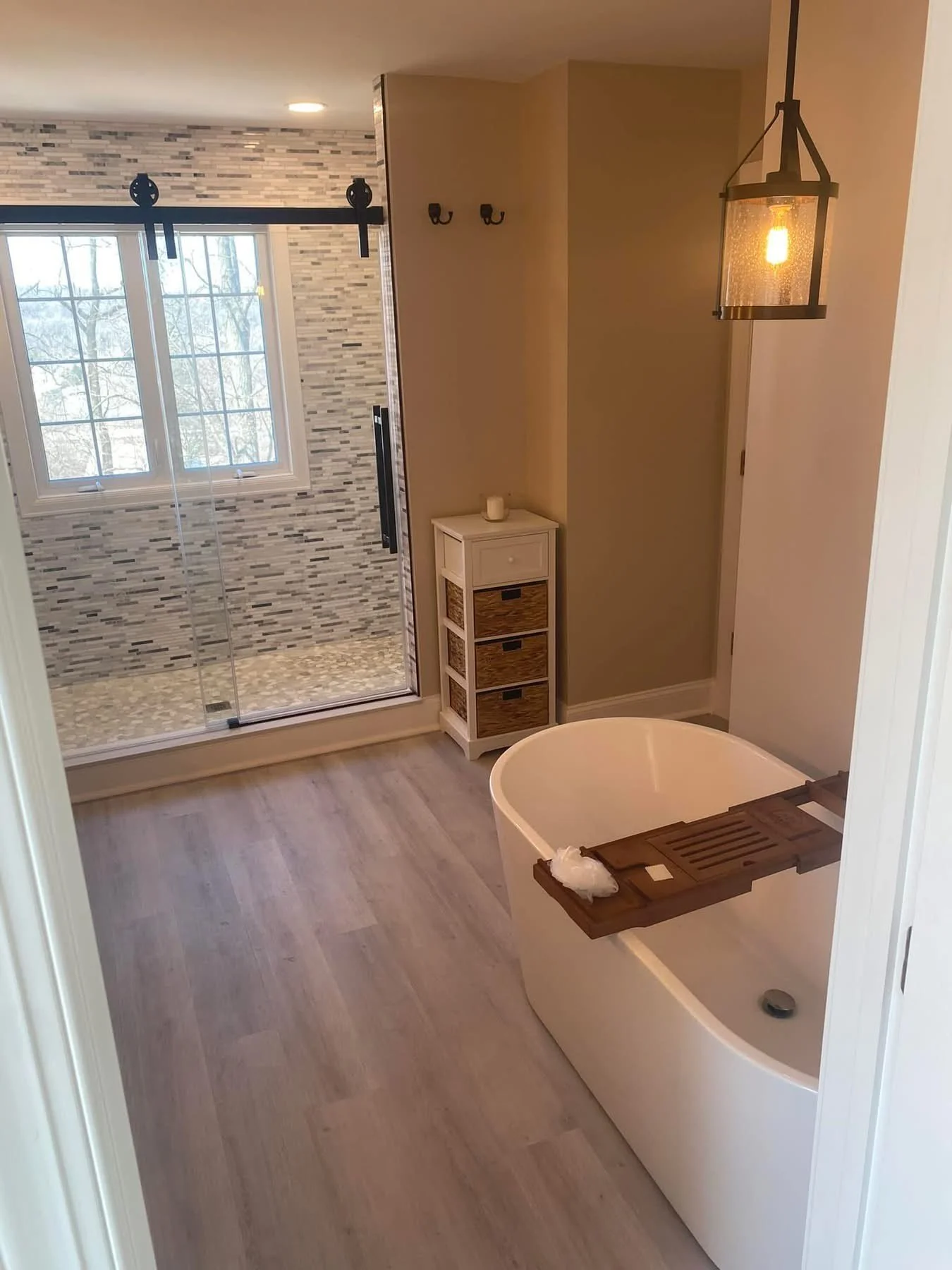 Bathroom with a freestanding bathtub, a small white storage cabinet beside the tub, a walk-in shower with stone tile walls and a glass door, a window overlooking trees, and a hanging light fixture.