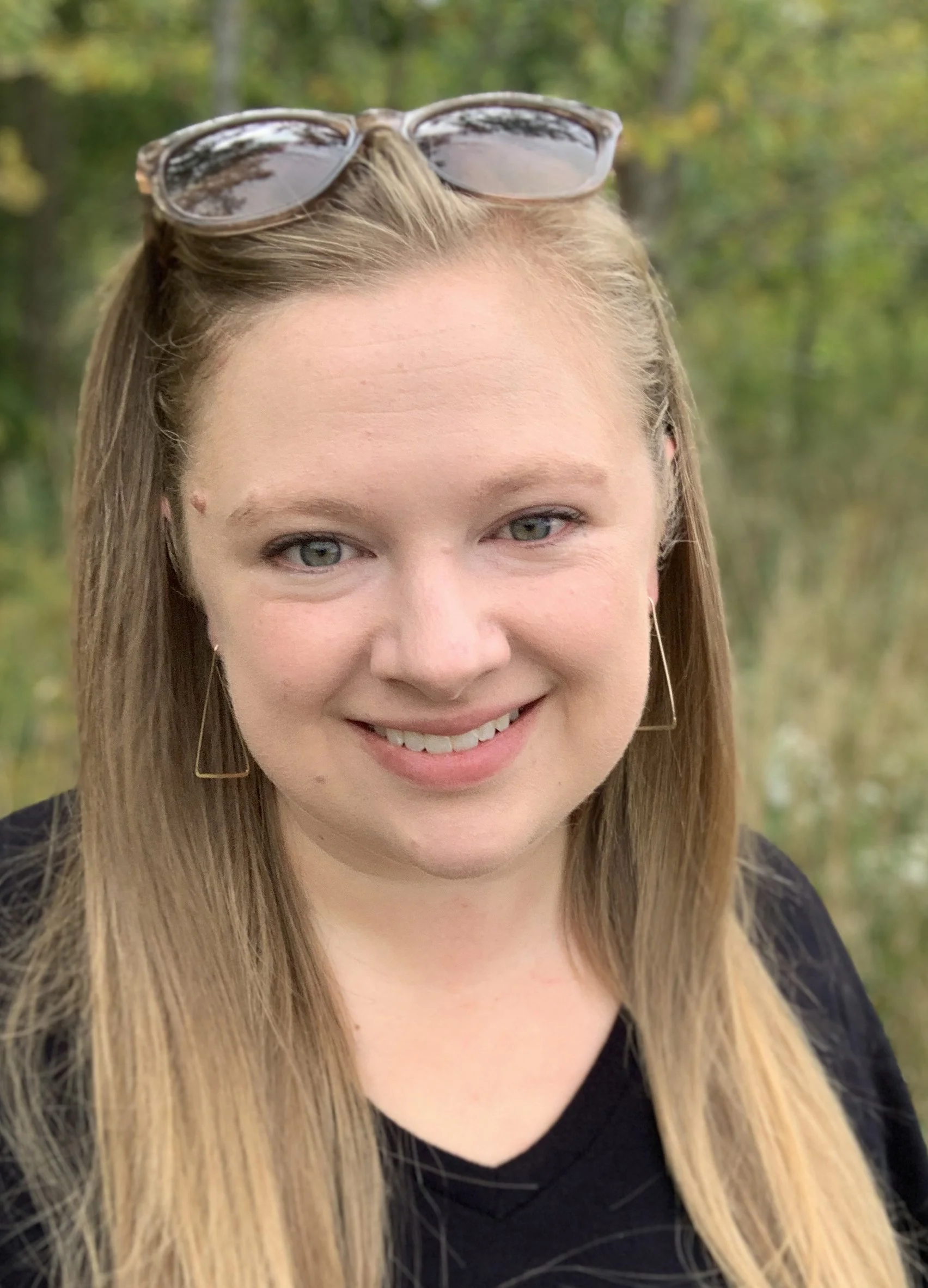 A woman with long blonde hair, wearing glasses on her head, gold triangle earrings, and a black top, smiling outdoors with green foliage in the background.