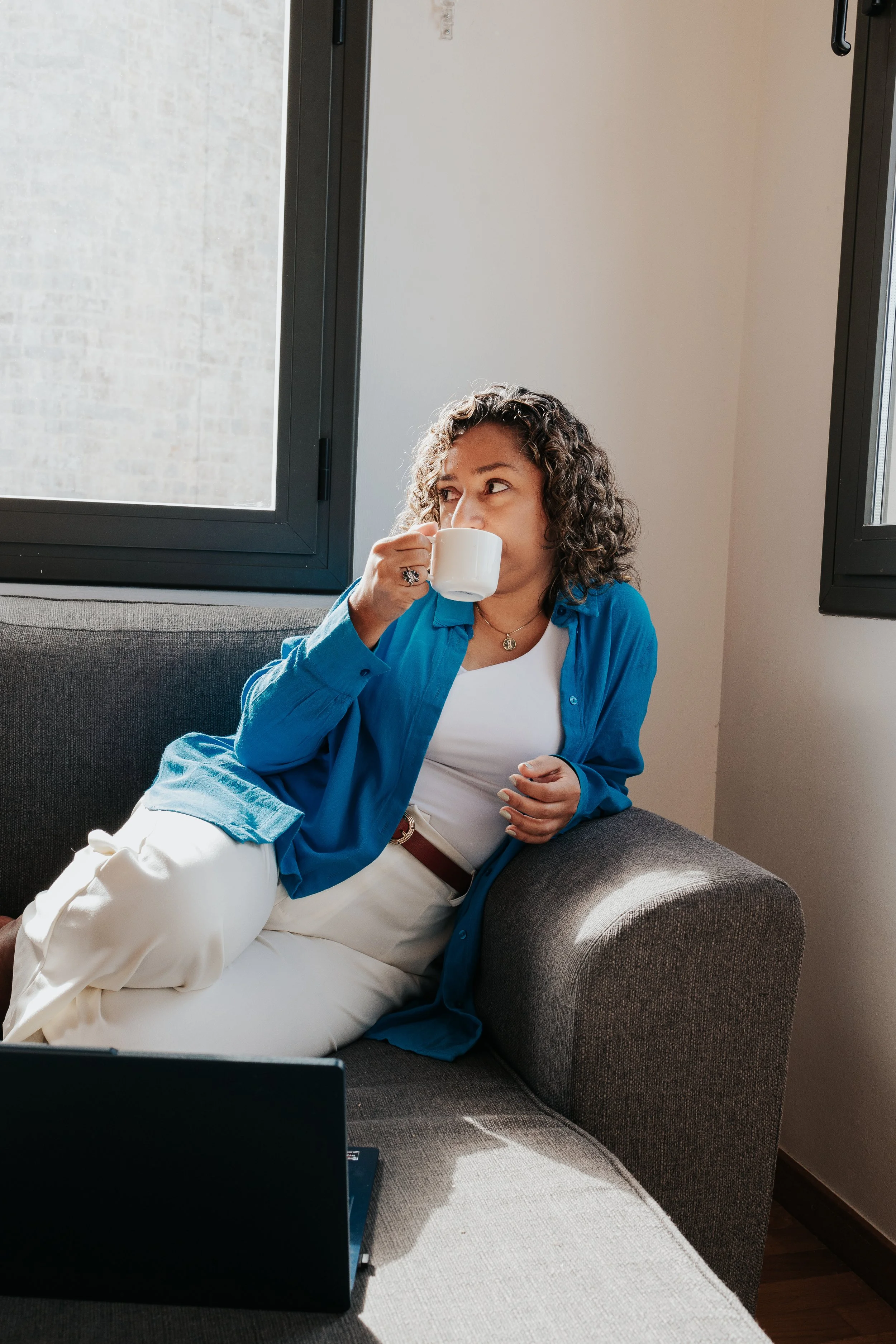 A woman with curly hair wearing a blue shirt and white pants sitting on a gray couch, drinking coffee near an open window.