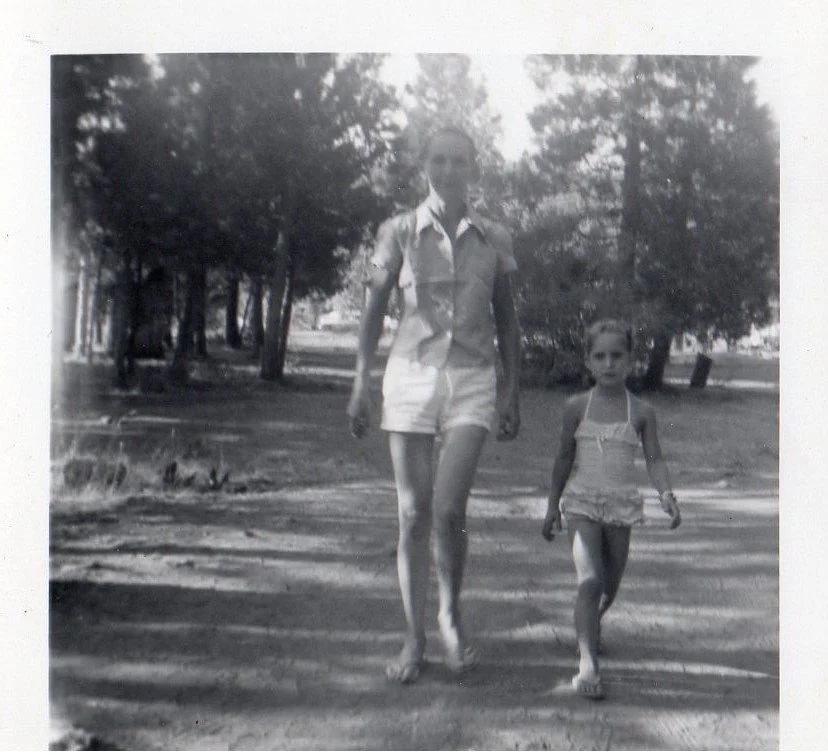 A black and white photo of a woman and a young girl walking outdoors on a dirt path surrounded by trees.