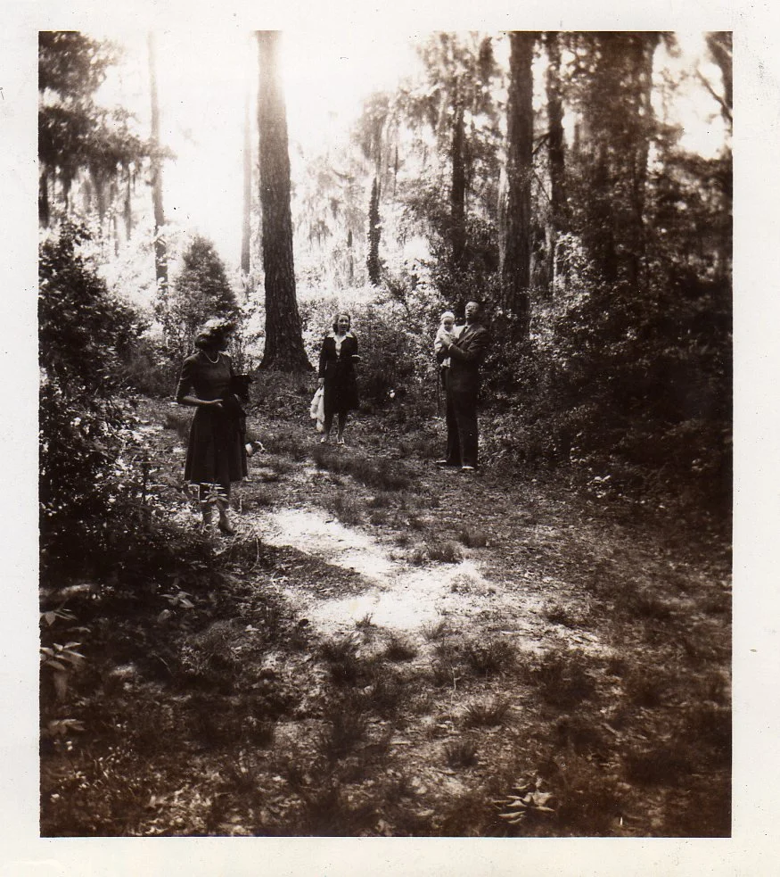A black-and-white photograph showing four people standing on a wooded trail in a forest, with tall trees and dense foliage around them.