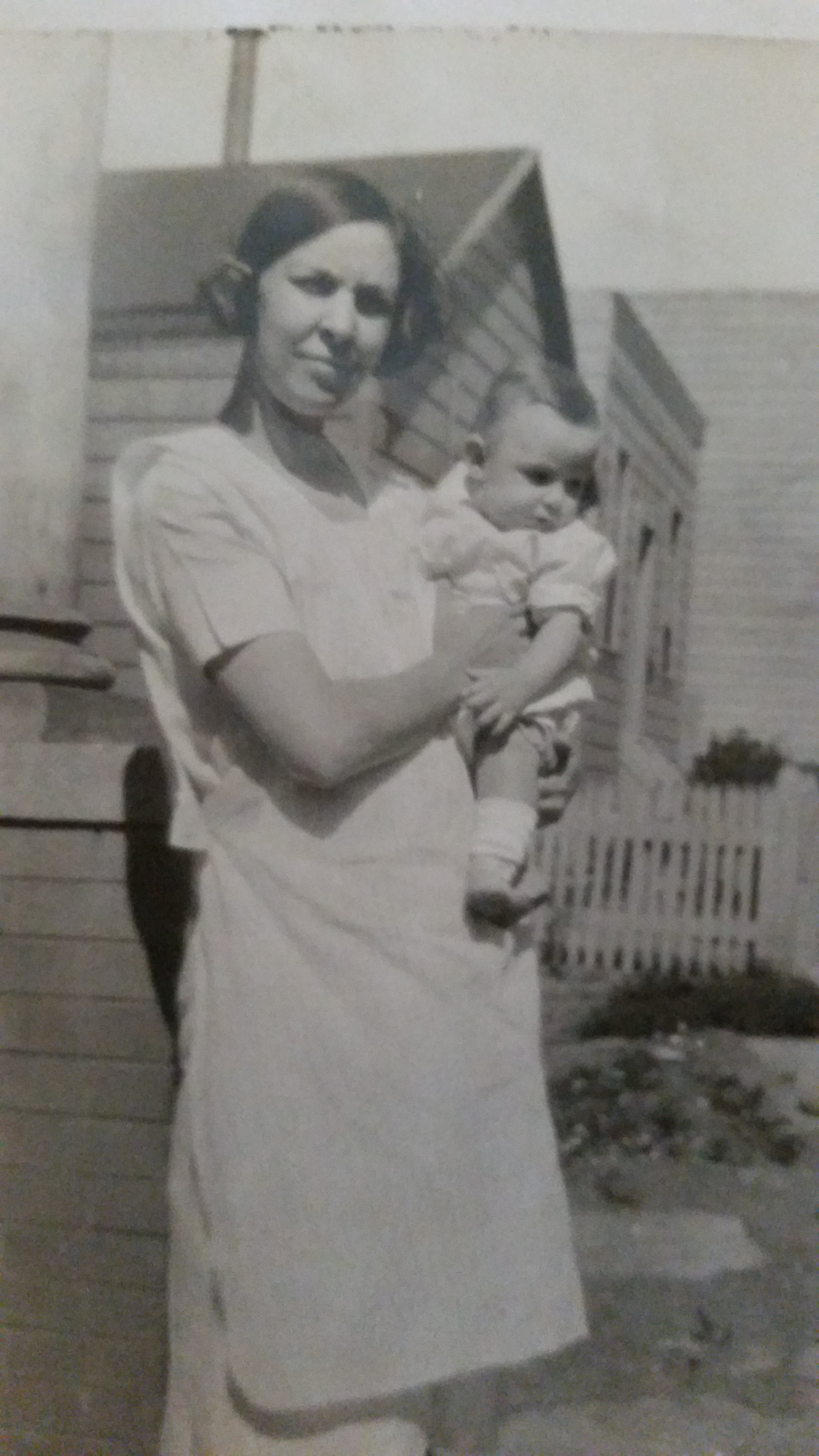 A woman in a dress holding a young child outdoors in front of a house with wooden siding, a picket fence, and a small garden.