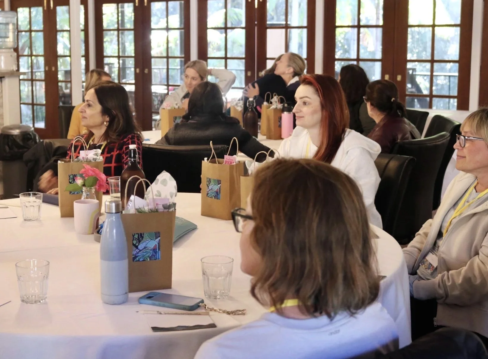 A group of women sitting around a large table in a bright room with large windows, participating in a workshop. The table has gift bags, water bottles, glasses, and a floral centerpiece.