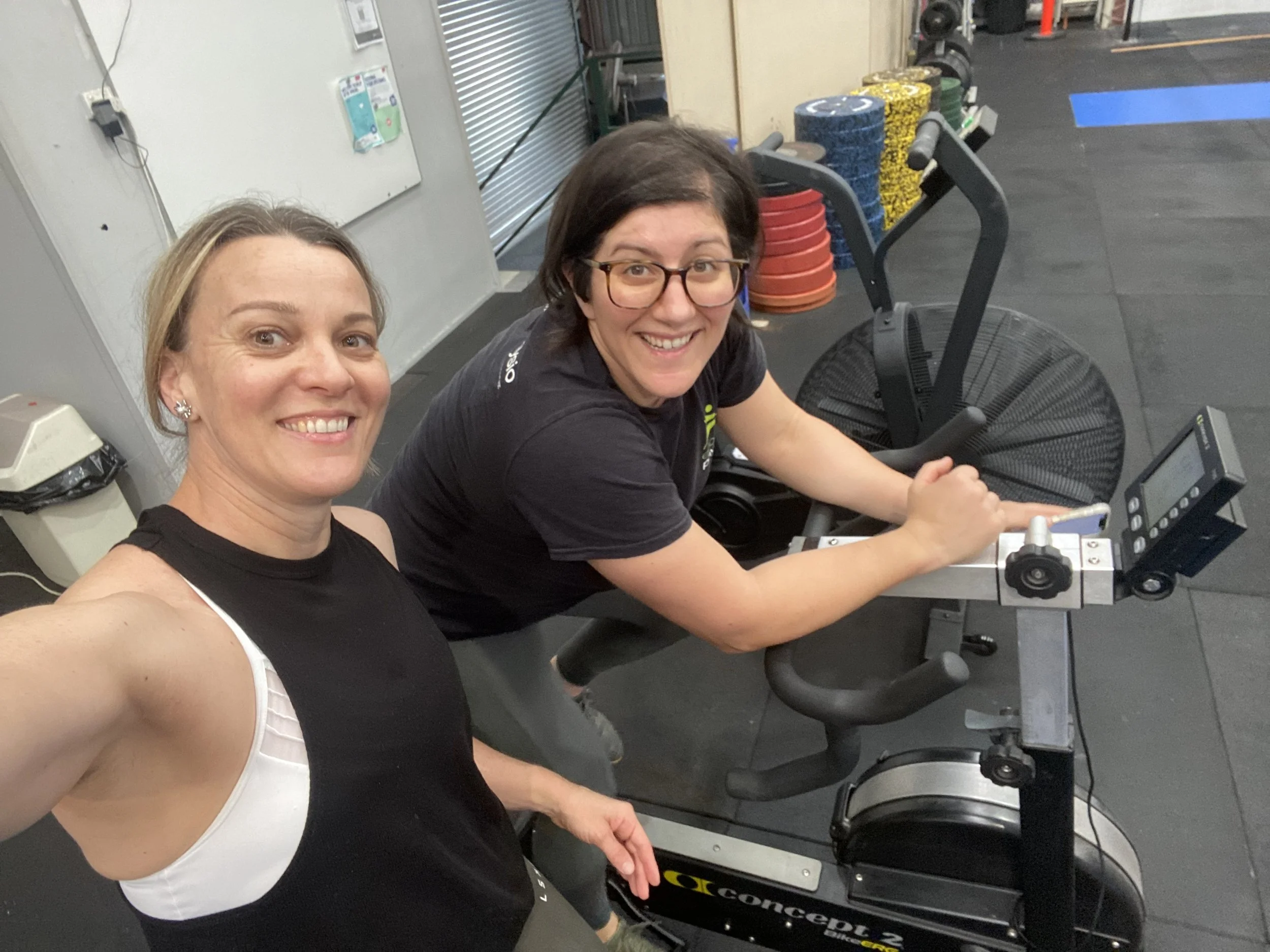 Two women smiling, one taking a selfie while the other is using a stationary exercise bike in a gym.