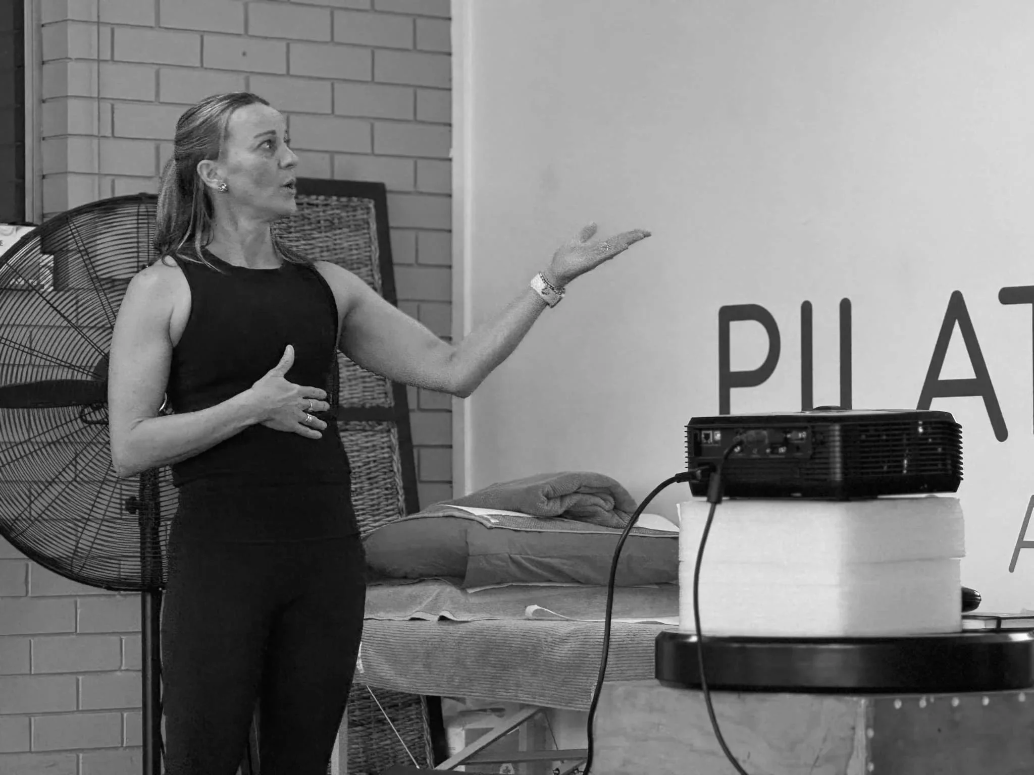 A woman in a sleeveless black top and black pants is gesturing while standing.