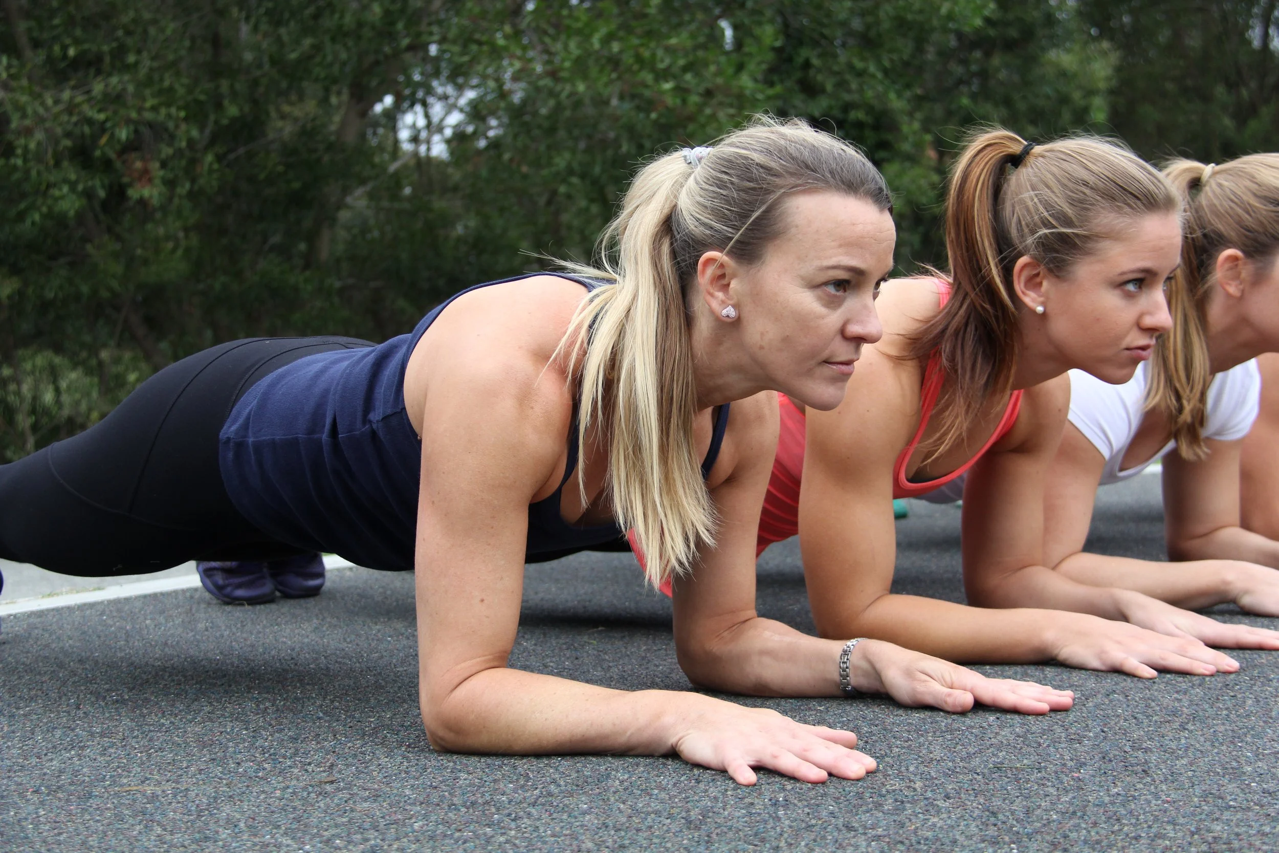 Women doing planks on a black outdoor surface surrounded by green trees.