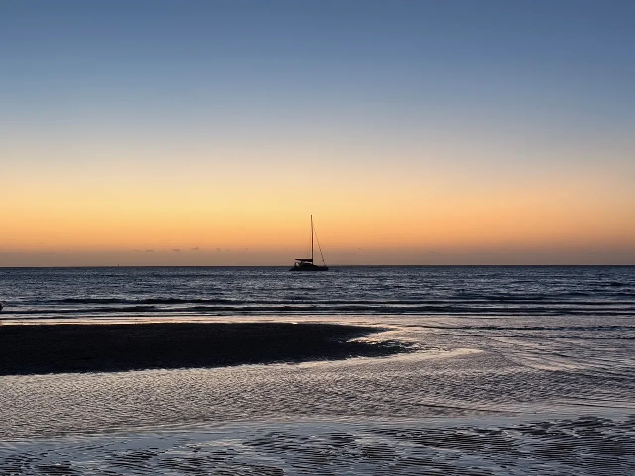 Sunset view of the ocean with a sailboat on the horizon, sandy beach in the foreground.