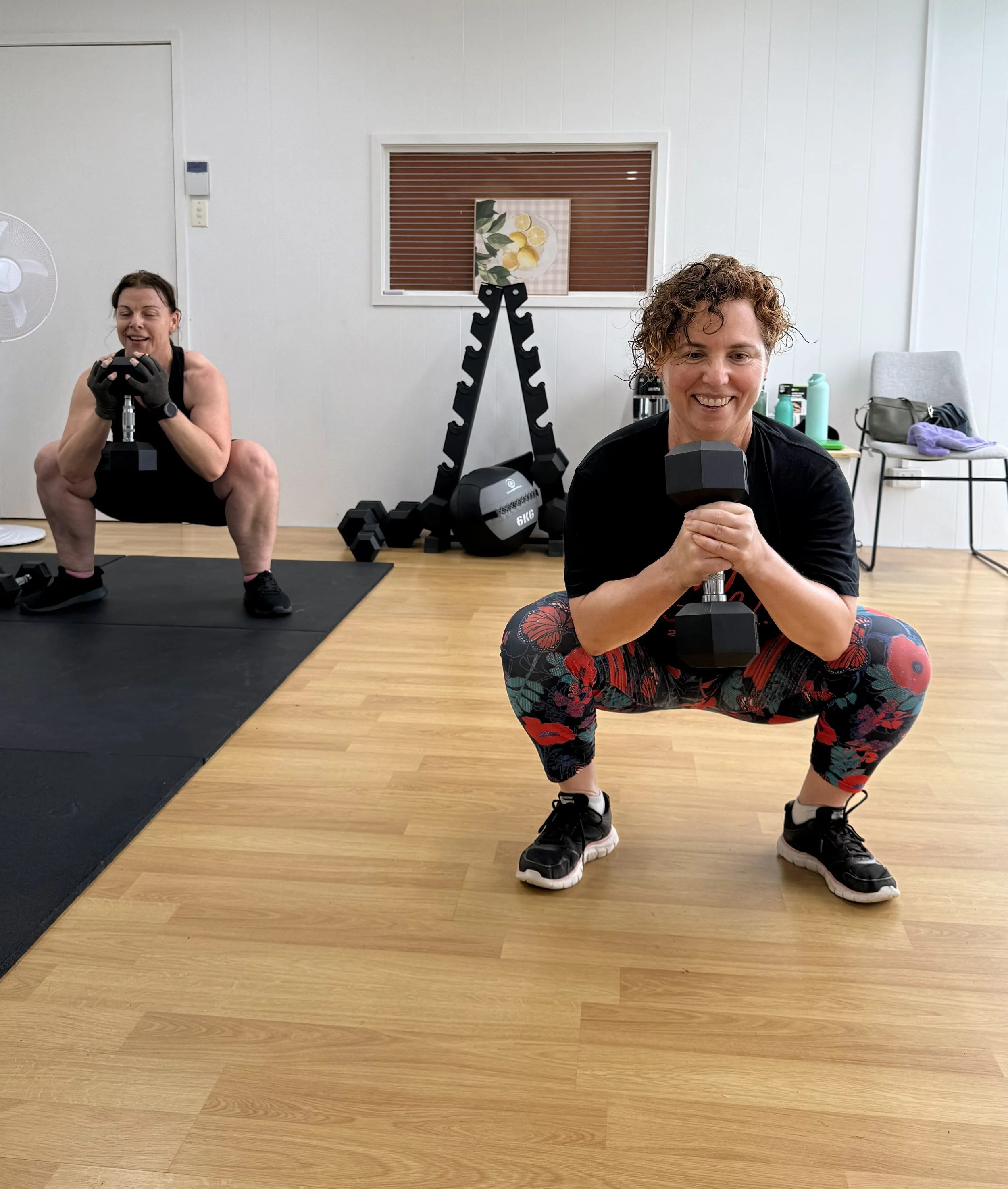 Two women exercising indoors, squatting with dumbbells, smiling at the camera. One woman in the foreground holding a dumbbell with both hands, wearing floral leggings and a black shirt. The other woman in the background also squatting, holding a dumbbell, with workout equipment and a picture of lemons on the wall behind them.