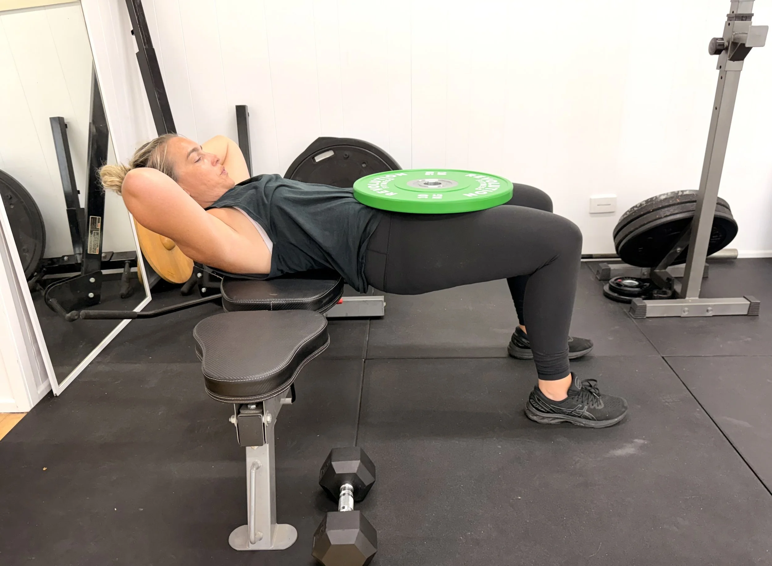 A woman lying on a workout bench with her hands behind her head, holding a yellow weight plate behind her neck, in a gym setting.