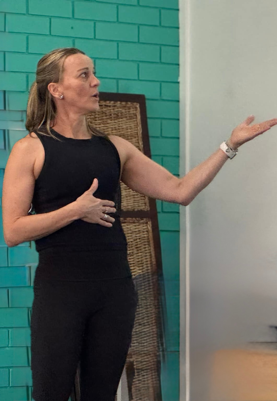 Woman giving a presentation or speech, gesturing with her hand, wearing a black sleeveless top and a smartwatch, standing in front of a teal brick wall.