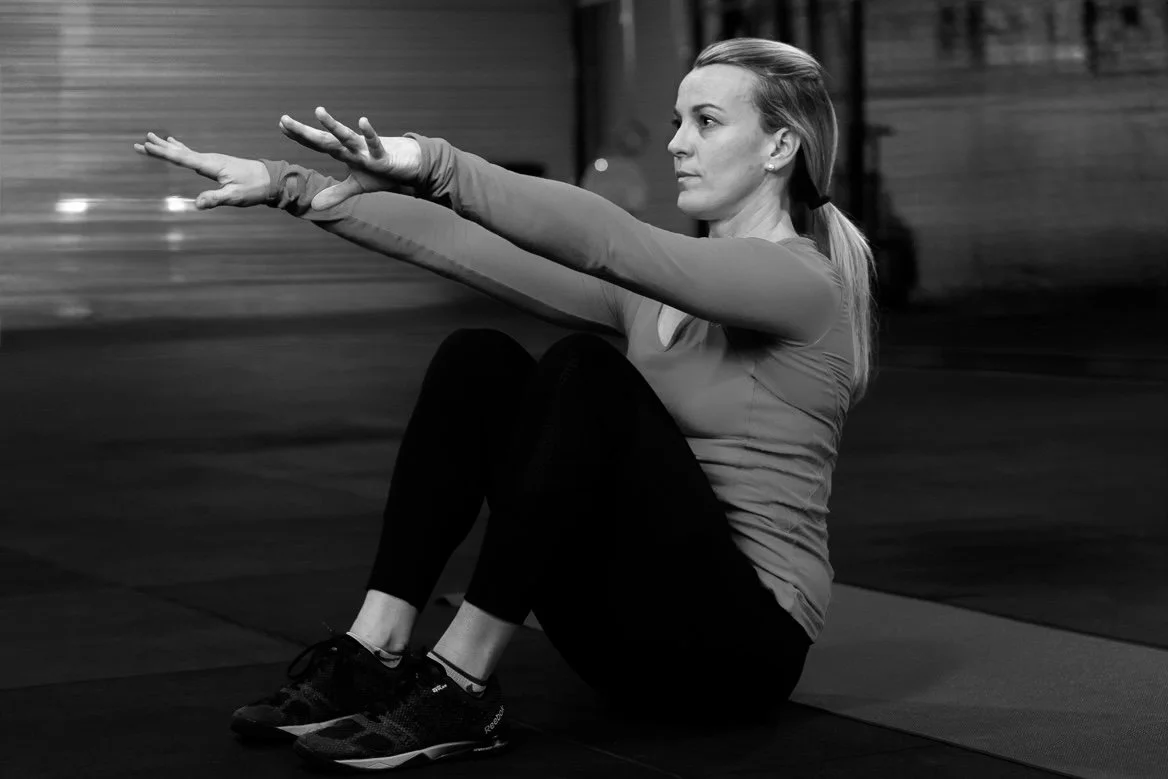 A woman sitting on a workout mat in a gym, stretching her arms forward with a focused expression.