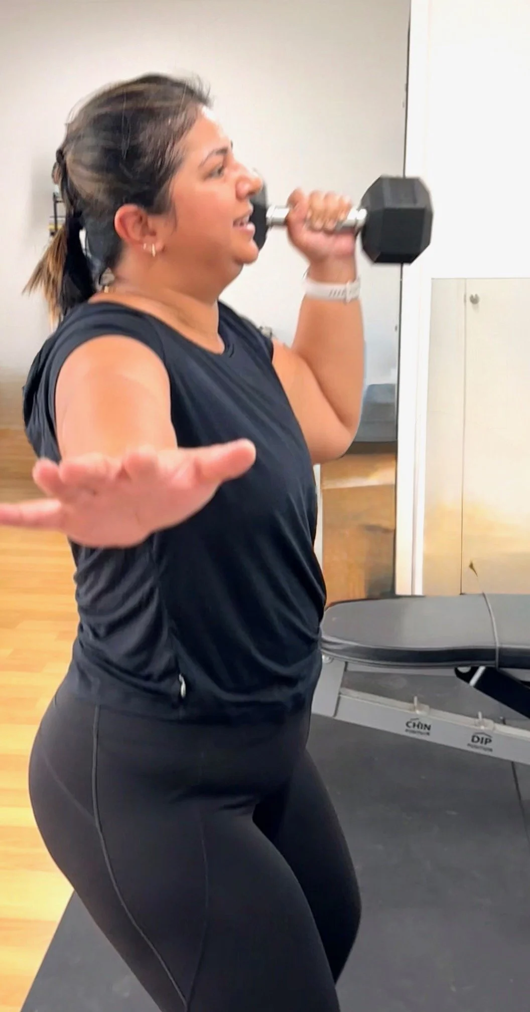 A woman working out with a dumbbell in a gym, holding it against her shoulder, with one arm extended and the other relaxed at her side.