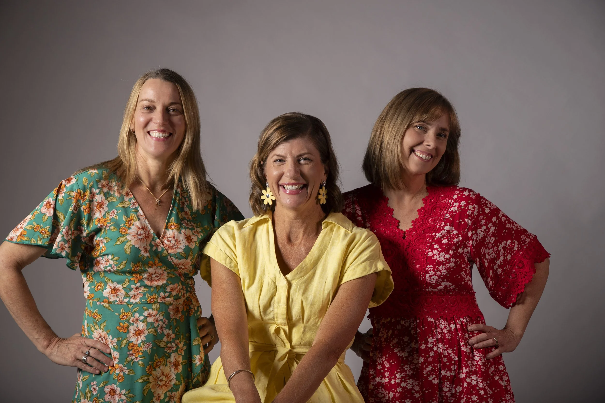 Three smiling women in colorful dresses posing together against a gray background.