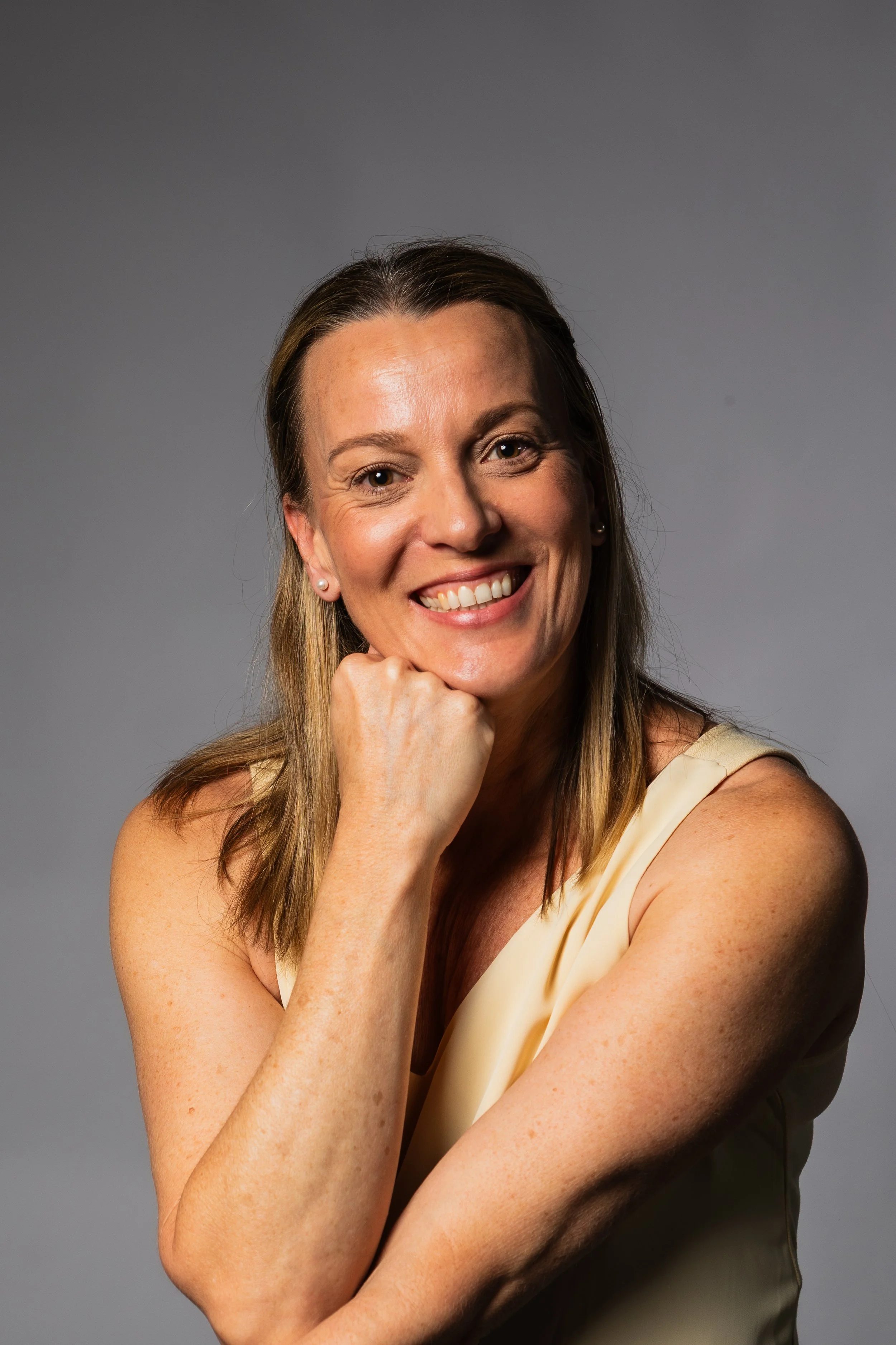 Smiling woman with blonde hair in a sleeveless cream top, resting her chin on her hand in a studio portrait against a gray background.