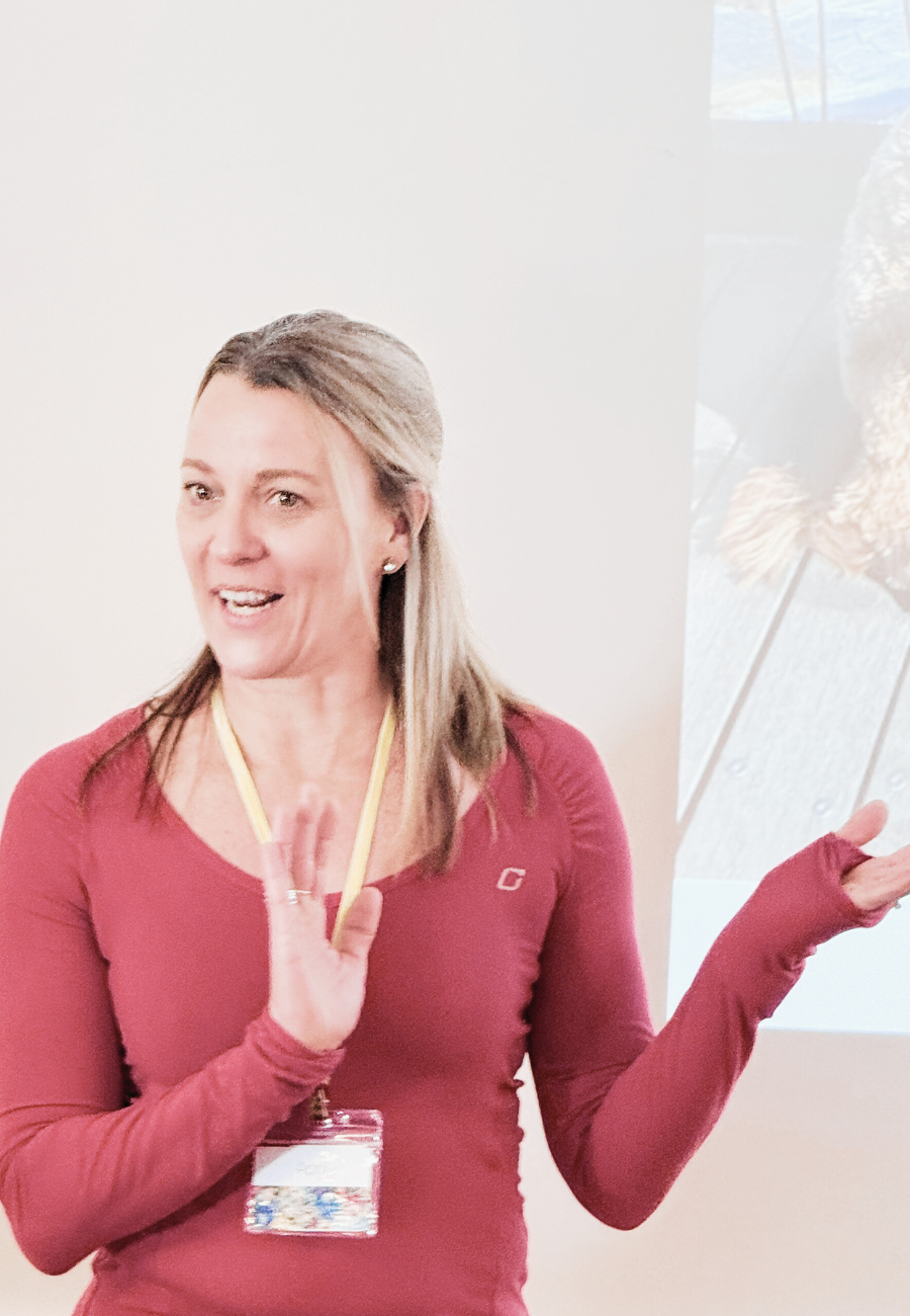 A woman wearing a red long sleeve shirt, standing and gesturing with her left hand, speaking during a presentation or discussion, with a projection of an outdoor scene.