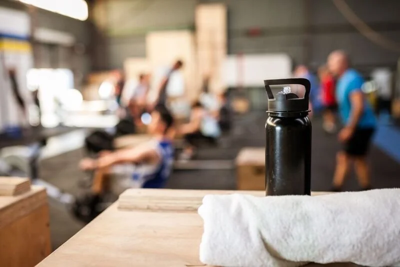 A black water bottle placed on a wooden table with a rolled-up white towel beside it. In the background, a group of people is working out in a gym with weights and exercise equipment.