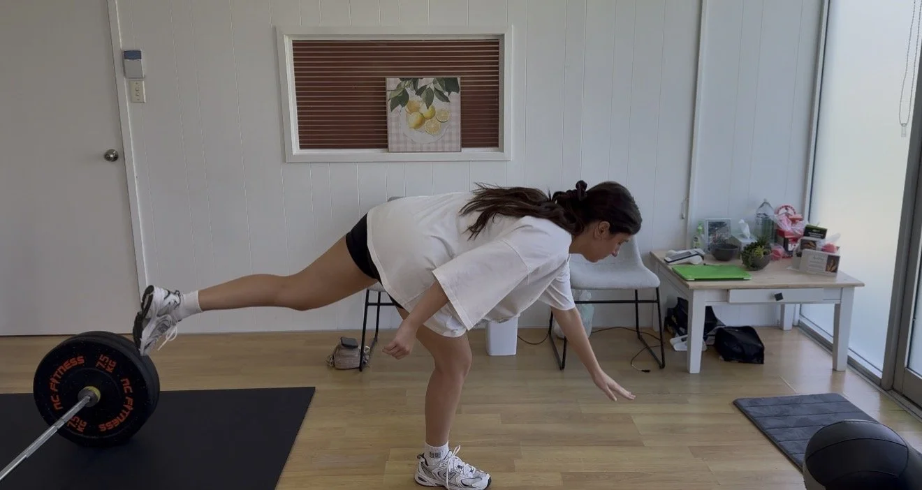 A woman is in a home gym balancing on one leg with one arm extended forward and the other behind, while holding a weight in her raised foot