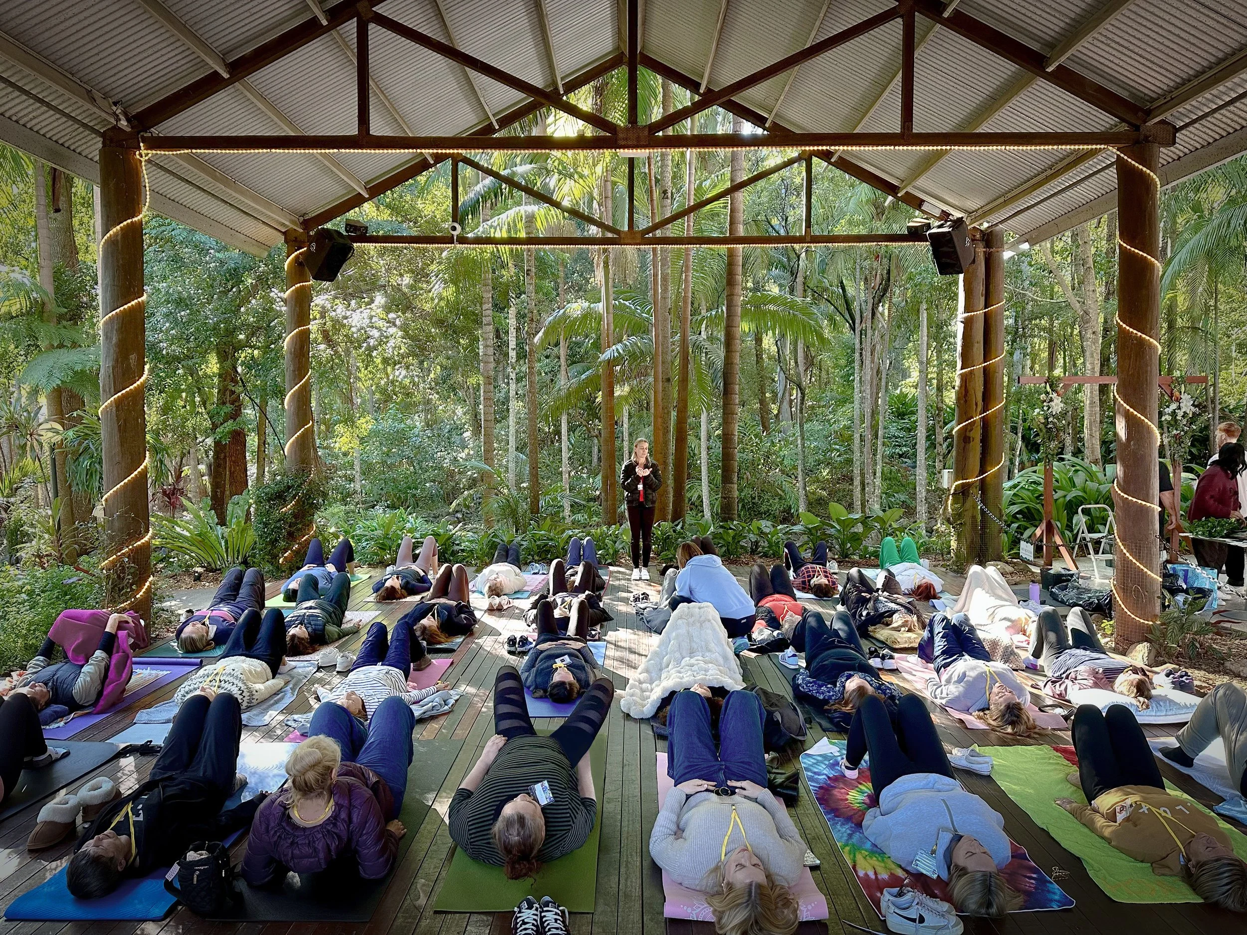 A large group of people participating in a yoga or meditation class in a forest pavilion with a metal roof, surrounded by trees and greenery.