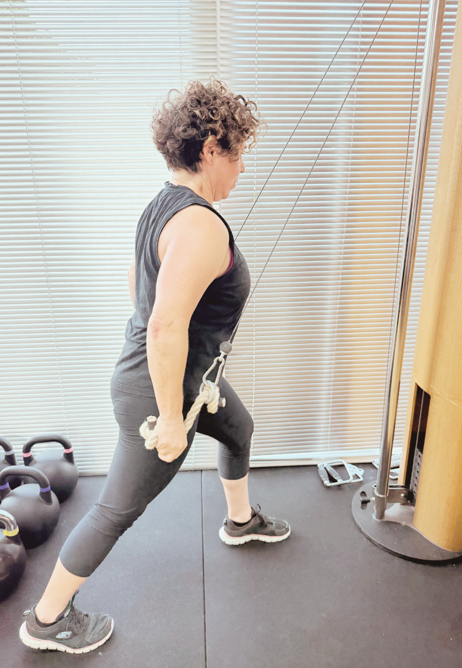 A woman exercising with a resistance band attached to a vertical pole, in a gym with kettlebells on the floor.