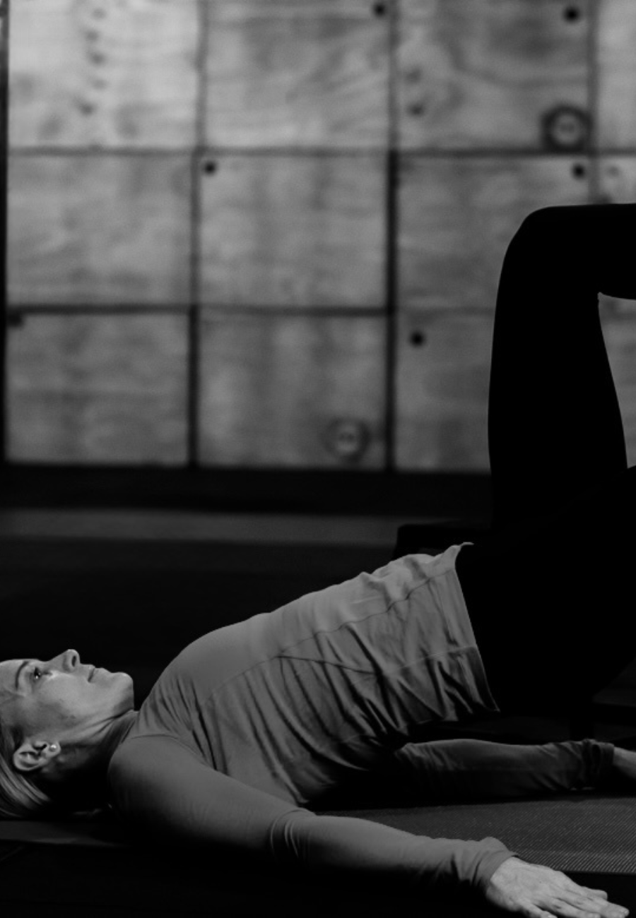 A woman is lying on her back on a yoga mat, performing a core exercise in a fitness studio with a wooden wall background.