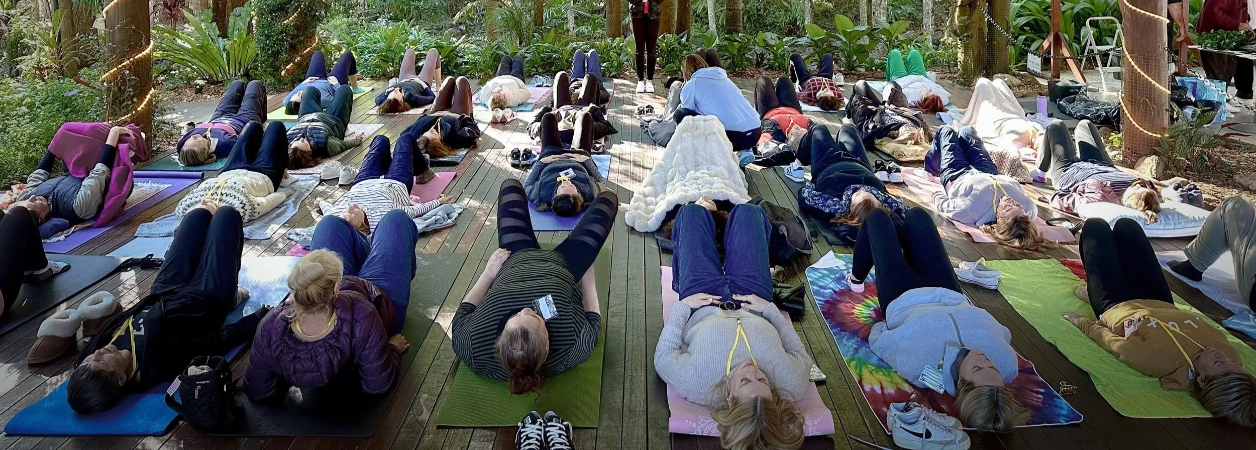 A group of people practicing yoga outdoors on wooden decking, lying on yoga mats amidst lush greenery.