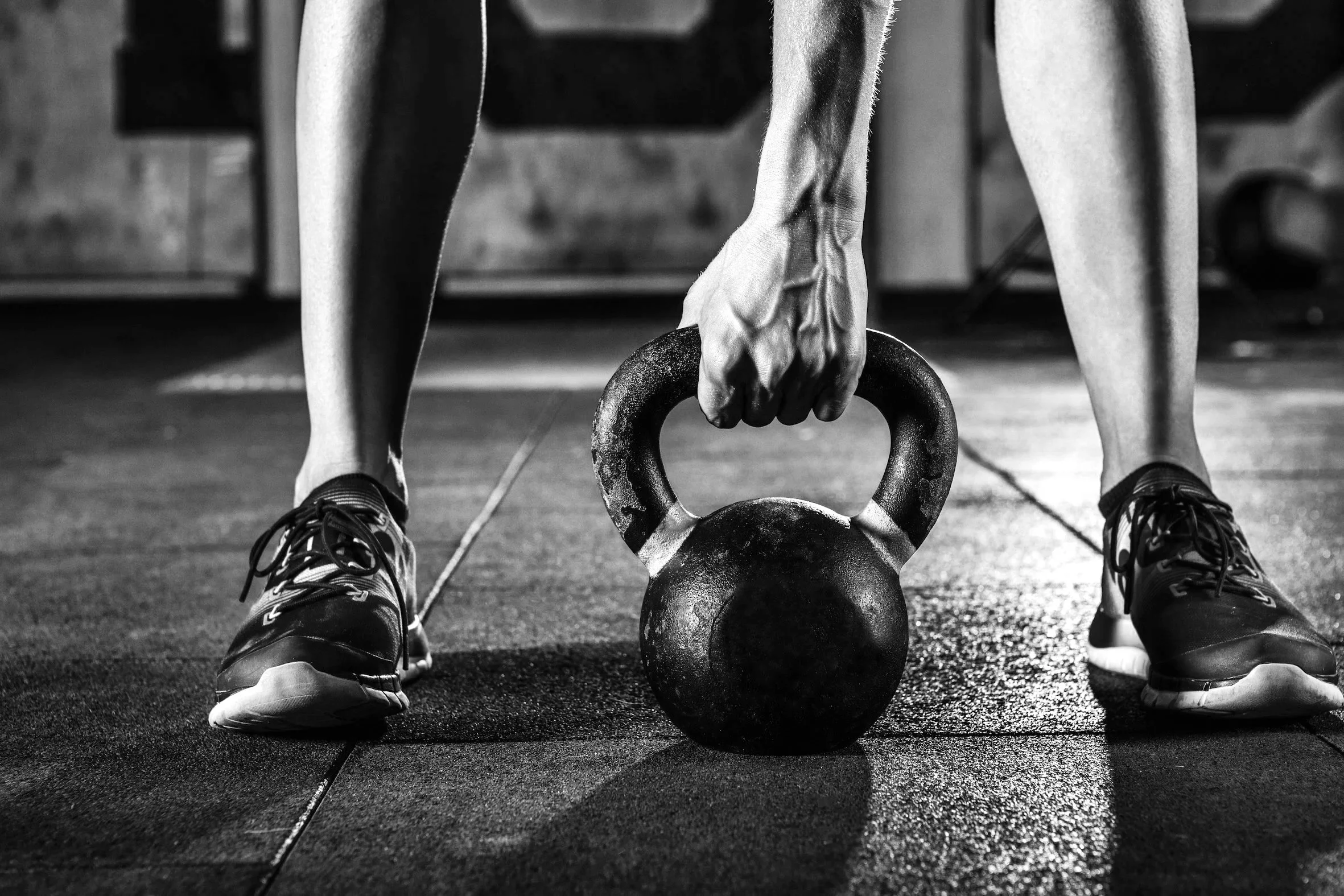 A person lifting a kettlebell in a gym, wearing athletic shoes, black and white photo.