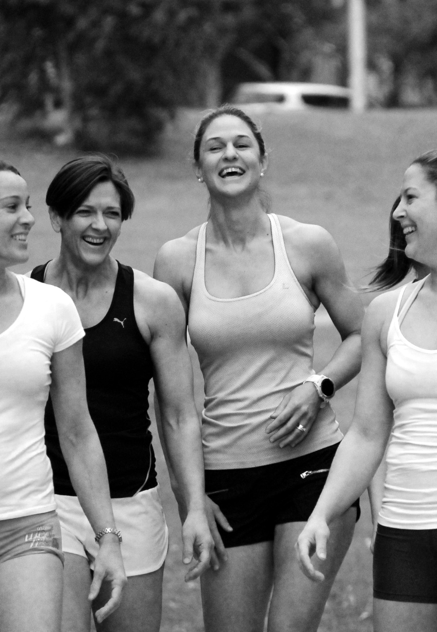 Four women in athletic clothing smiling and walking outdoors in a park.