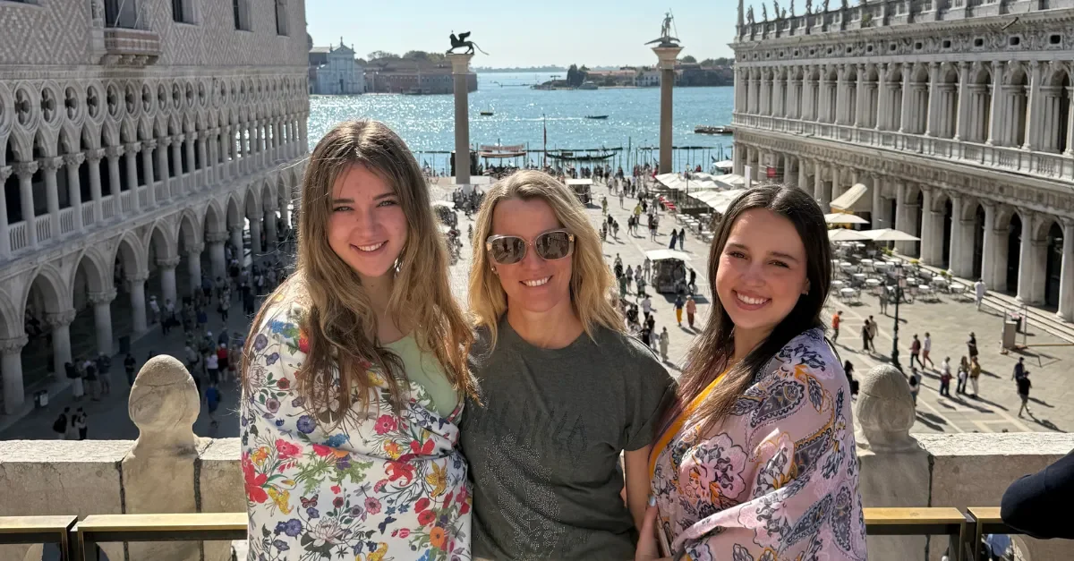 Three women smiling in front of a historic cityscape with water, boats, and statues in Venice, Italy.