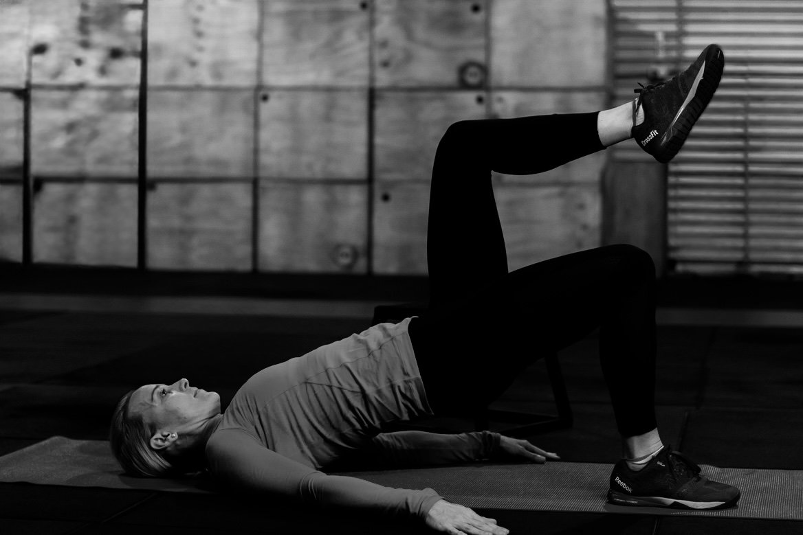 A woman exercises on a yoga mat in a gym using a leg lift while lying on her back.