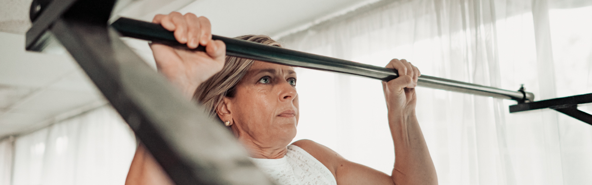 A middle-aged woman lifting a barbell during a workout in a bright gym.