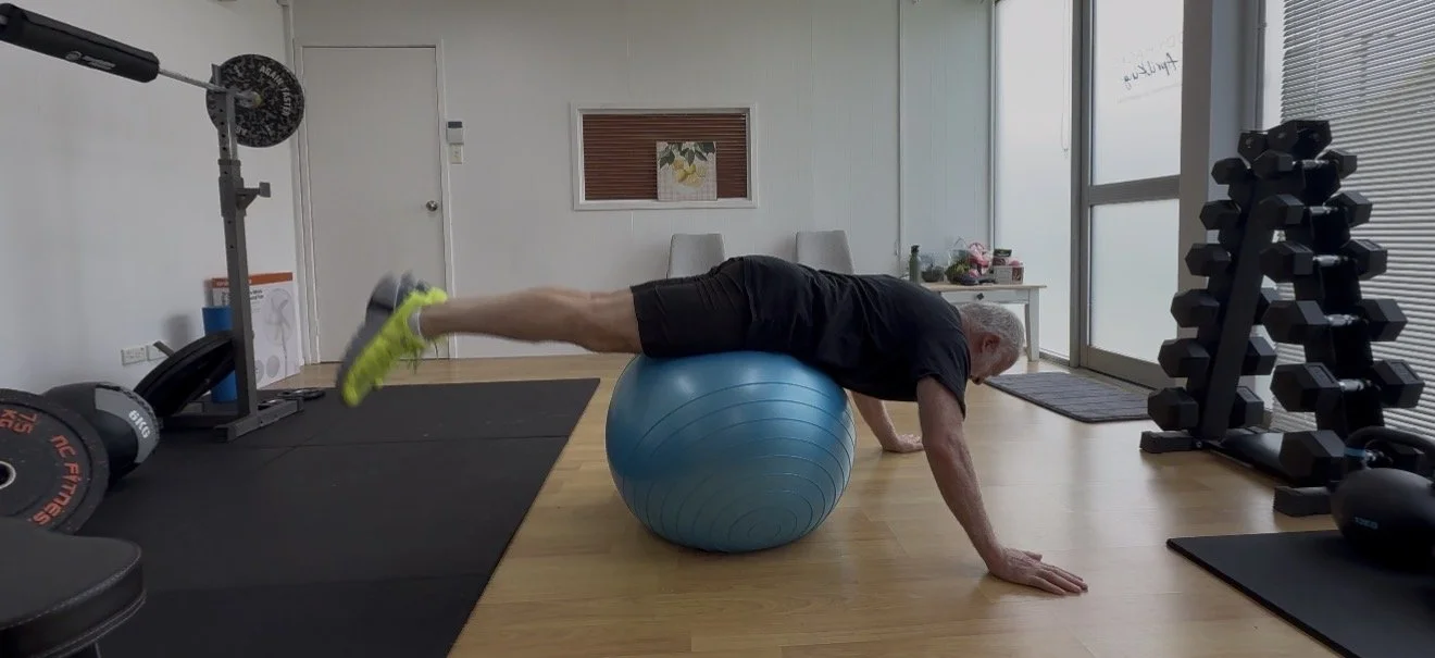 Senior man exercising on a fitness ball in a gym, reaching forward with his hands on the floor.