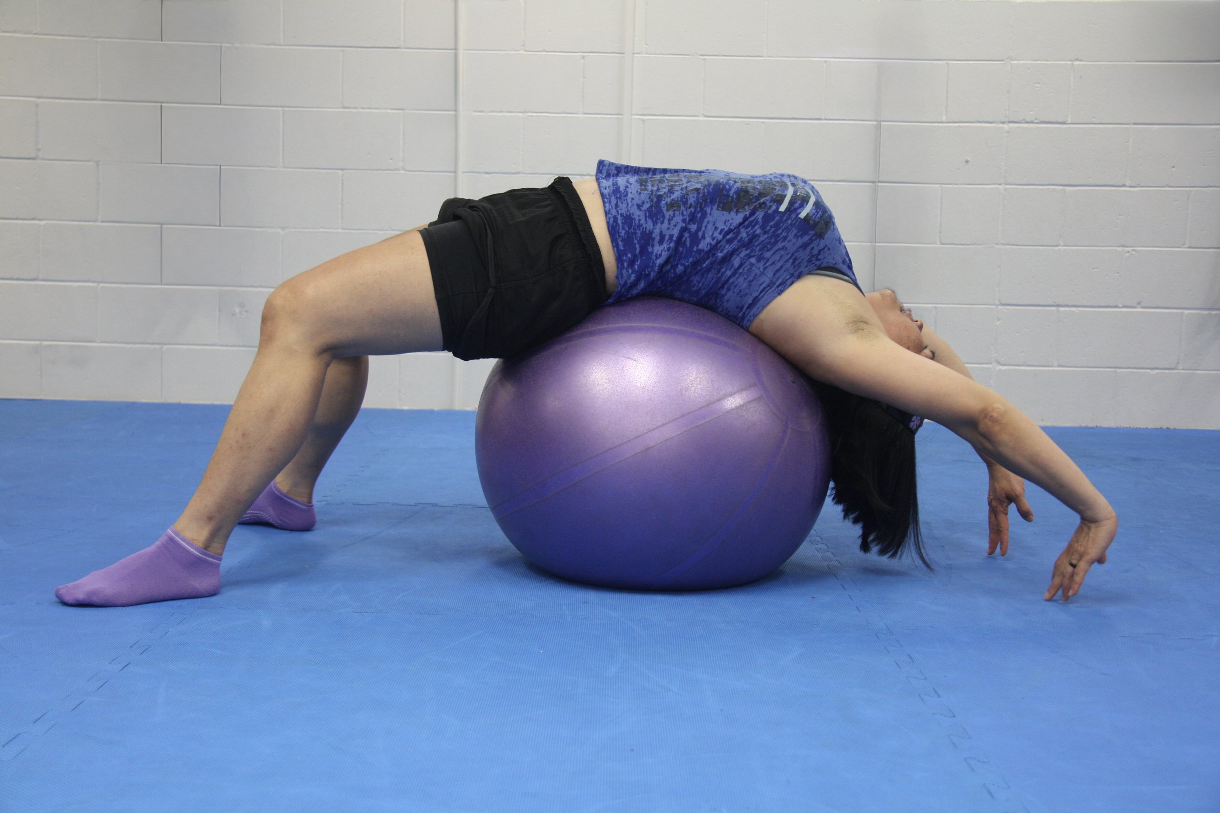 A woman performing an exercise on a purple stability ball in a gym, lying on her back with her hips elevated, arms extended overhead, and legs bent.