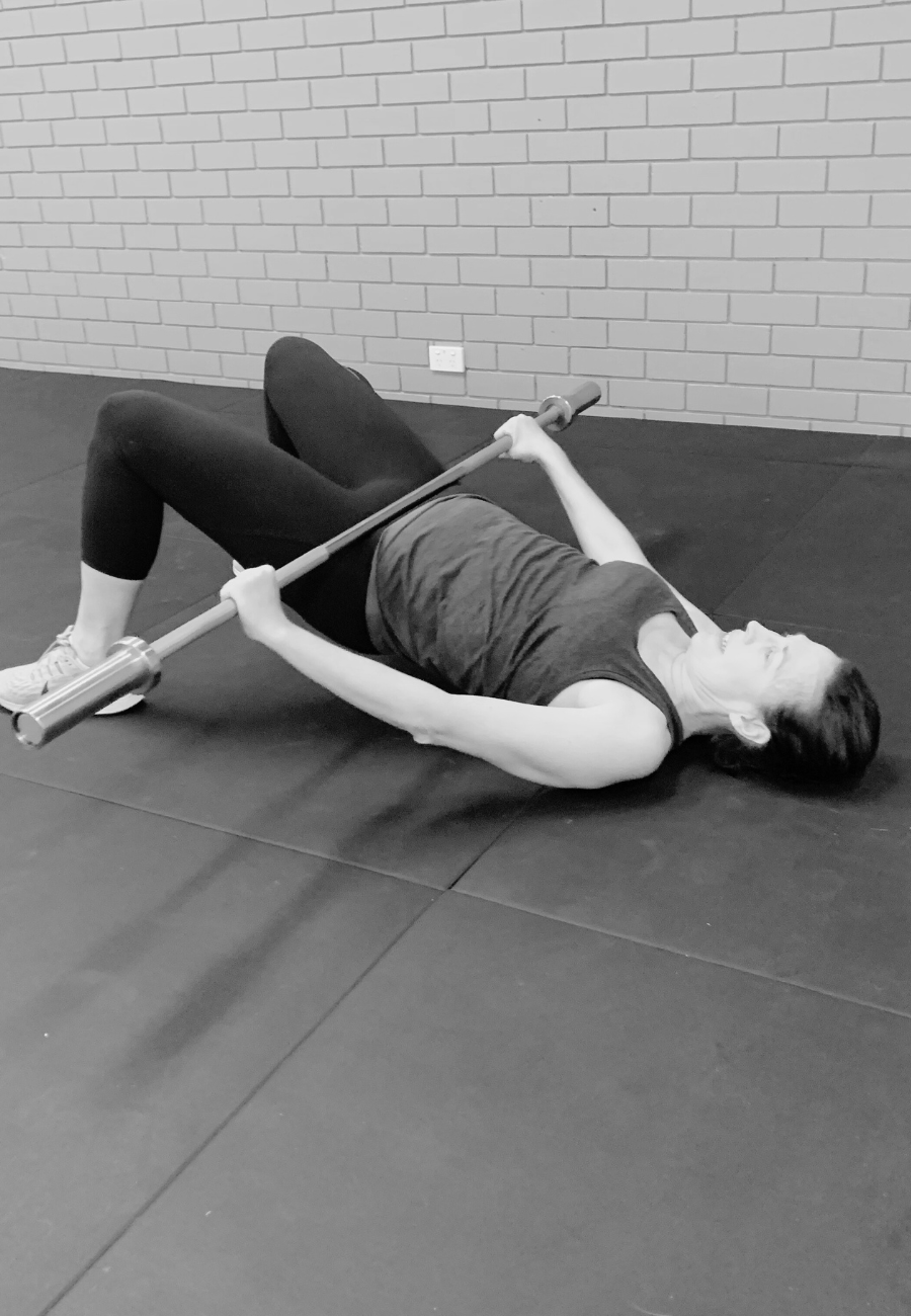 A woman lying on the floor performing a bridge exercise while holding a barbell.