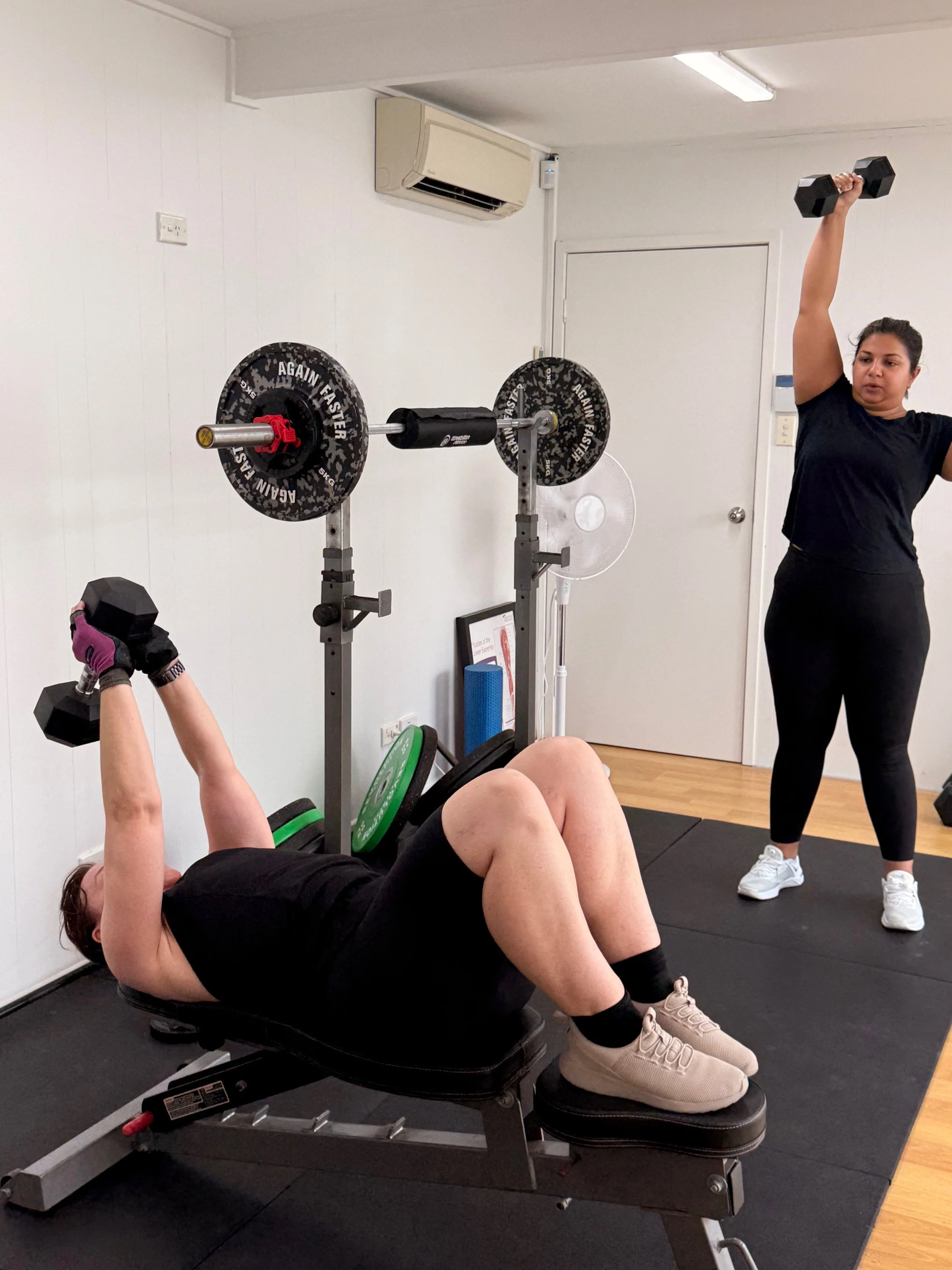 Two women working out in a gym, one lying on a bench with dumbbells and the other standing and holding a dumbbell overhead.