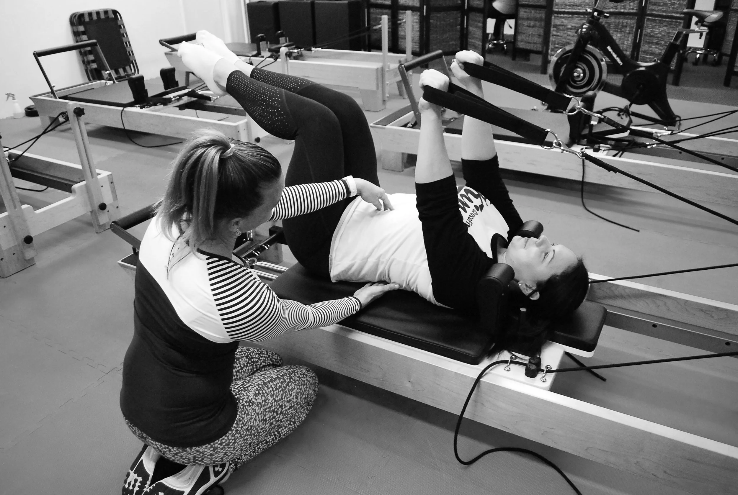 A woman is lying on a Pilates reformer machine, performing an exercise with straps held in her hands, while another woman assists her by holding her waist.