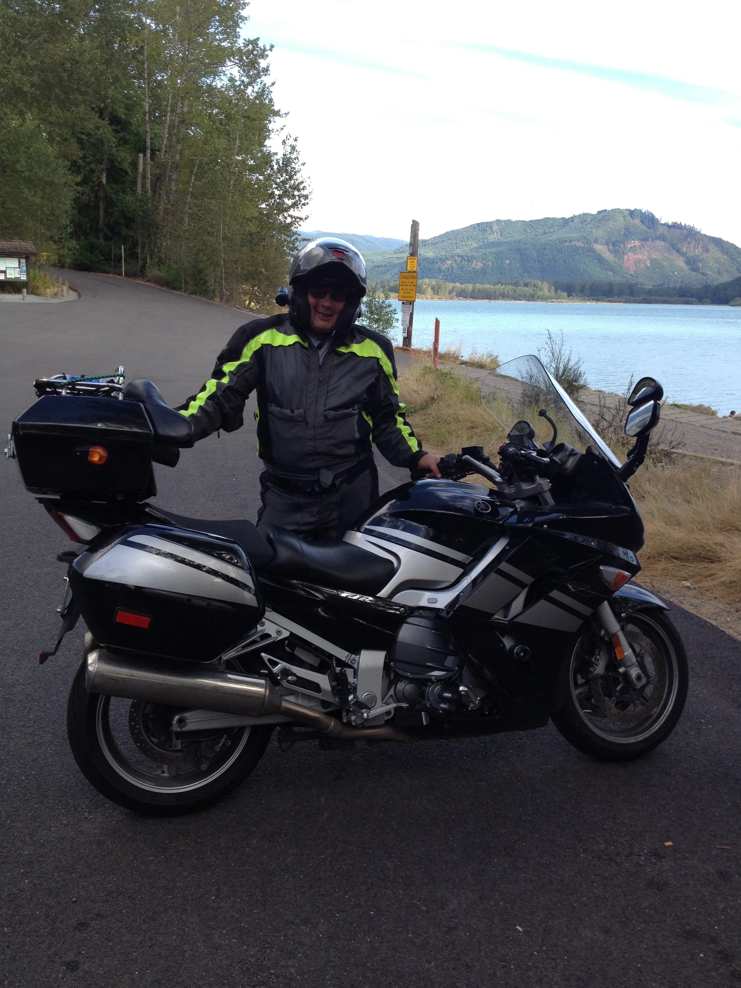 Person in riding gear and helmet standing next to a black and white touring motorcycle by a lake with forested hills in the background.