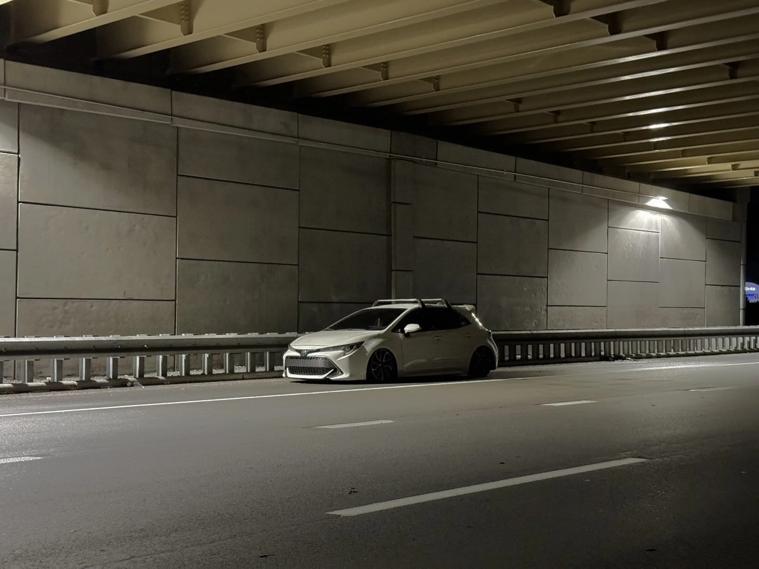 A white compact car parked on the side of a highway at night under an overpass.