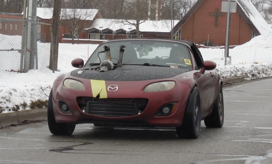 A red Mazda convertible car with a damaged yellow stripe on the front, parked in a lot with snow and a church in the background.