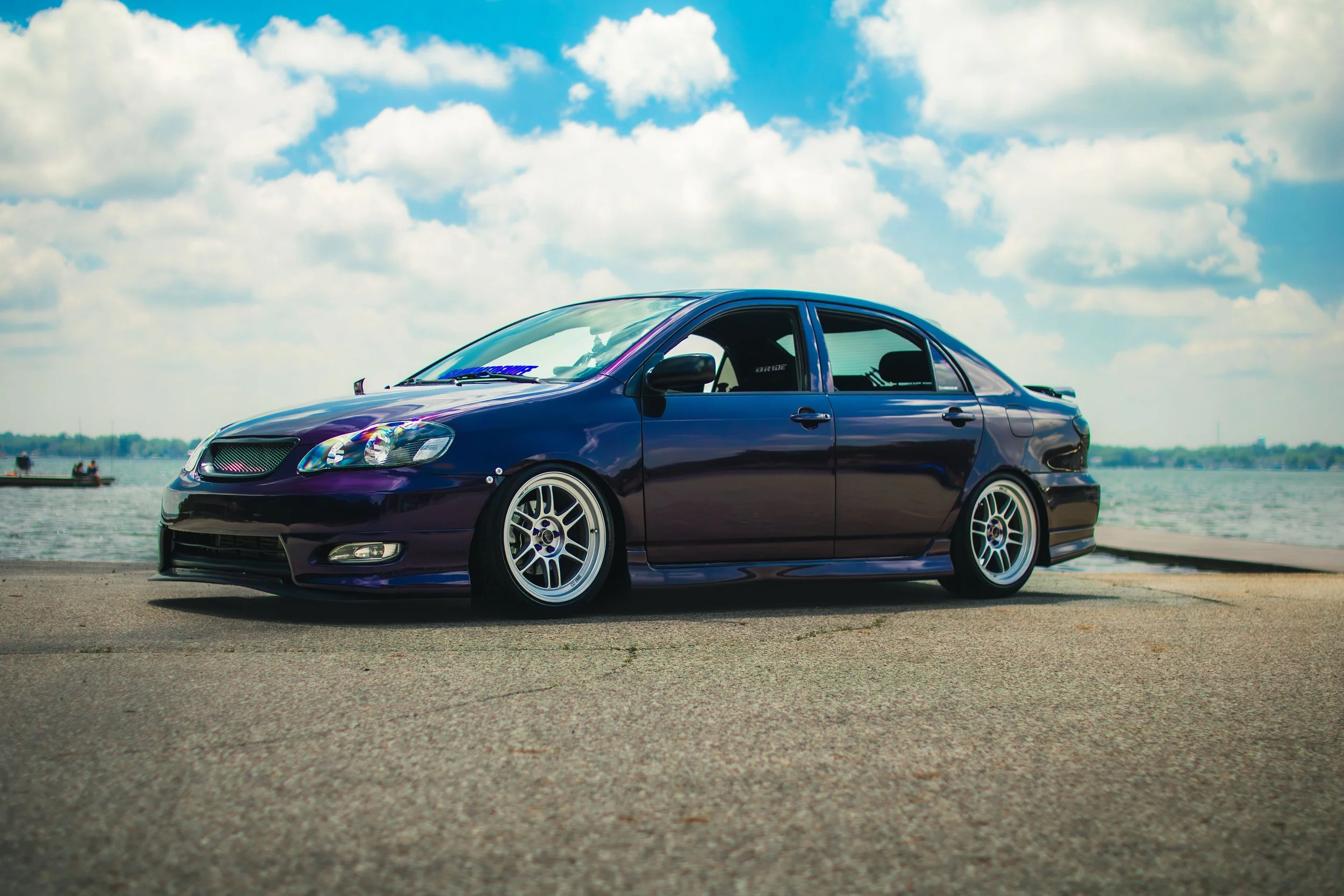 Dark purple modified sedan car parked on pavement near water with boats and clouds in the sky.