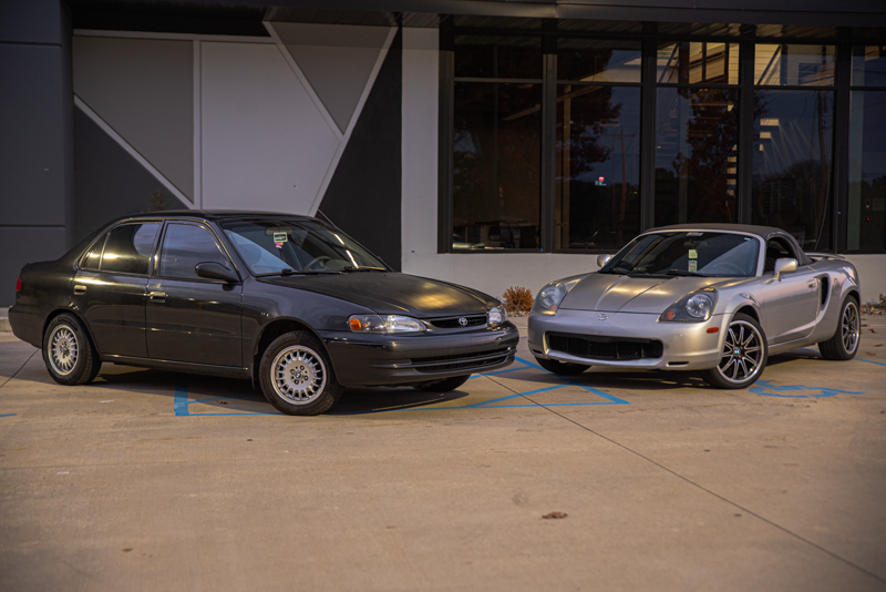 A black sedan and a silver sports car are parked side by side in a parking lot near a modern building with large glass windows at dusk.